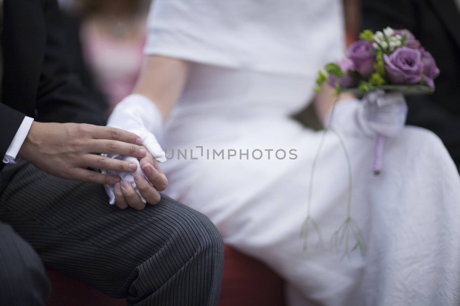 Color artistic digital photo of bridegroom in dark suit and white shirt with cufflinks in church religious wedding marriage ceremony holding hands with the bride in white wedding bridal dress. Shallow depth of with background out of focus. 