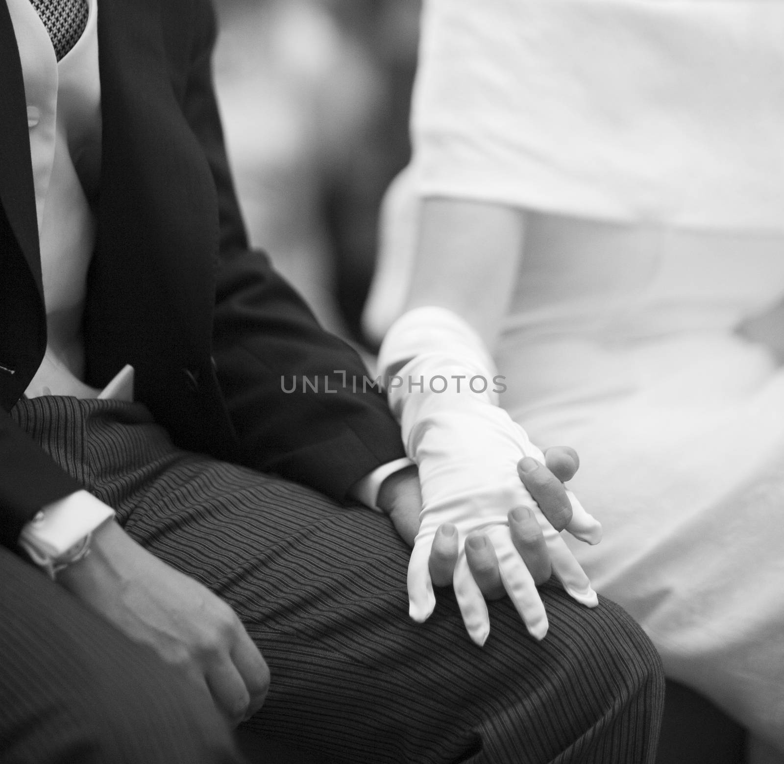 Black and white artistic digital photo of bridegroom in dark suit and white shirt in church religious wedding marriage ceremony holding hands with the bride in white long wedding bridal dress. Shallow depth of with background out of focus. 