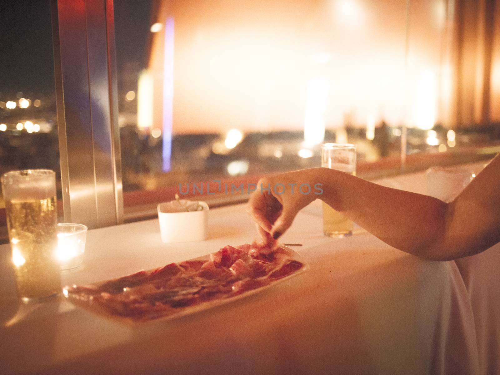 Color artistic digital rectangular horizontal photo of the hand of a woman taking piece of Spanish hand cut Iberian cured acorn fed black foot pig ham "Jamon iberico de pura bellota de pata negra" from plate of prosciutto style cured ham in Spanish wedding banquet marriage buffet party at night with window and city out of focus in defocused background. 