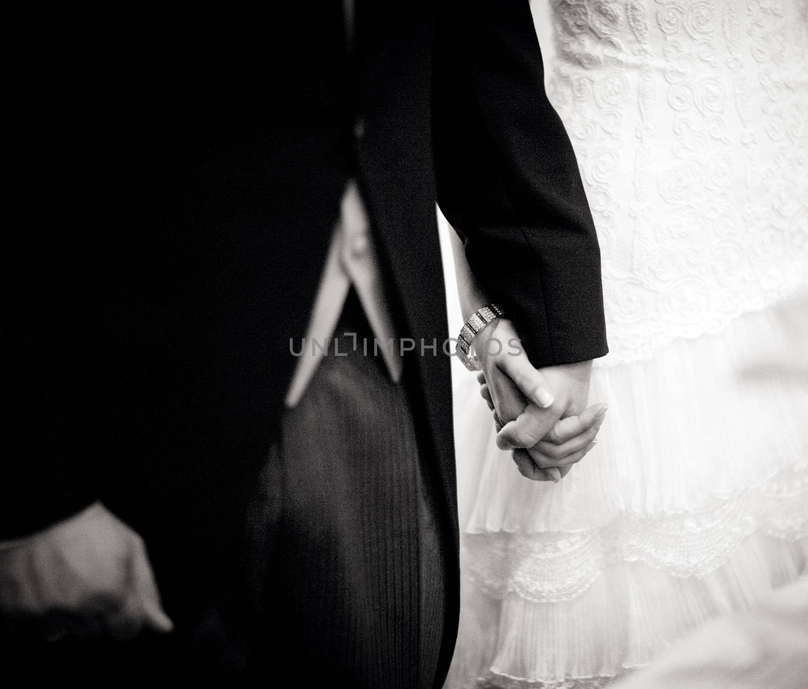 Black and white artistic digital rectangular horizontal photo of the bride wearing white wedding dress holding hands with the bridegroom wearing long jacket dark morning suit during church wedding marriage ceremony in Madrid Spain. Shallow depth of on foreground with background out of focus. 