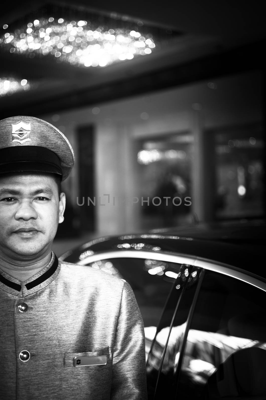 Bangkok, Thailand - November 29, 2013: Parking attendant standing outside luxury five star gran luxe hotel in Bangkok dowtown in Thailand wearing silk Thai uniform. 

Black and white high contrast digital photo of a parking attendant in posh silk Thai uniform and cap outside a luxury five star hotel in Bangkok Thailand with hotel and luxury car behind out of focus. Shallow dof depth of focus and bokeh effect. 
