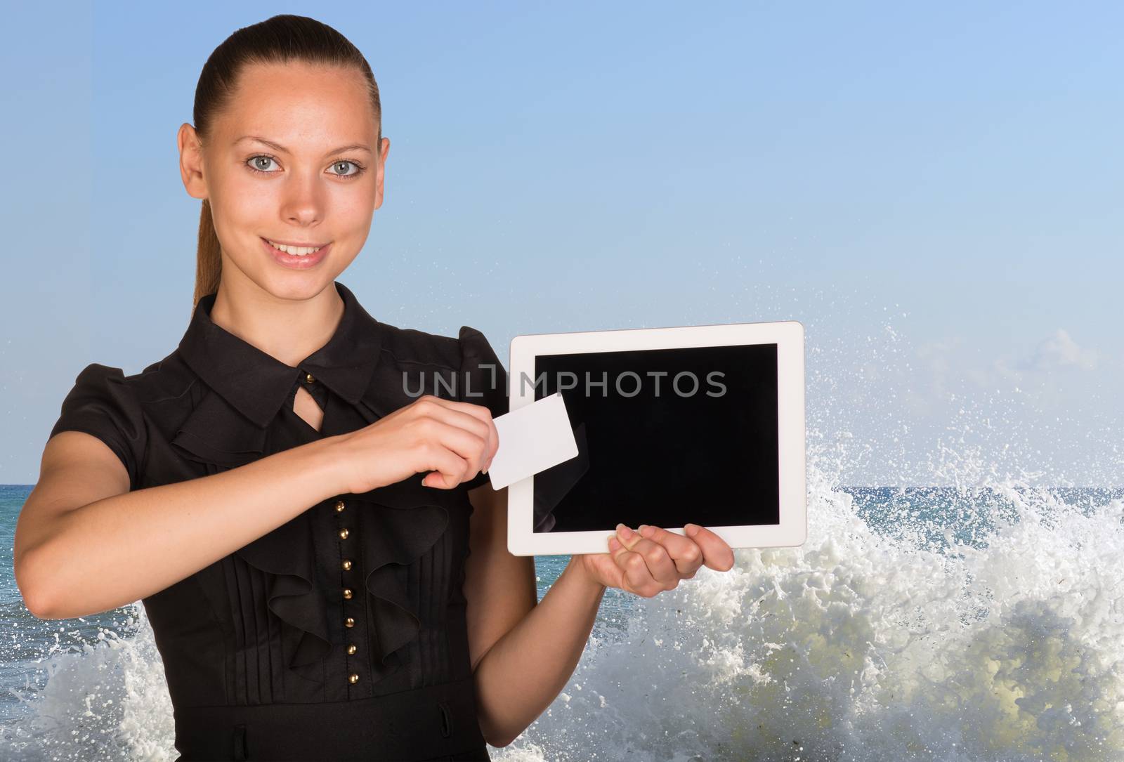 Beautiful businesswoman holding blank tablet PC and blank business card in front of PC screen. Seascape whith tidal waves as backdrop