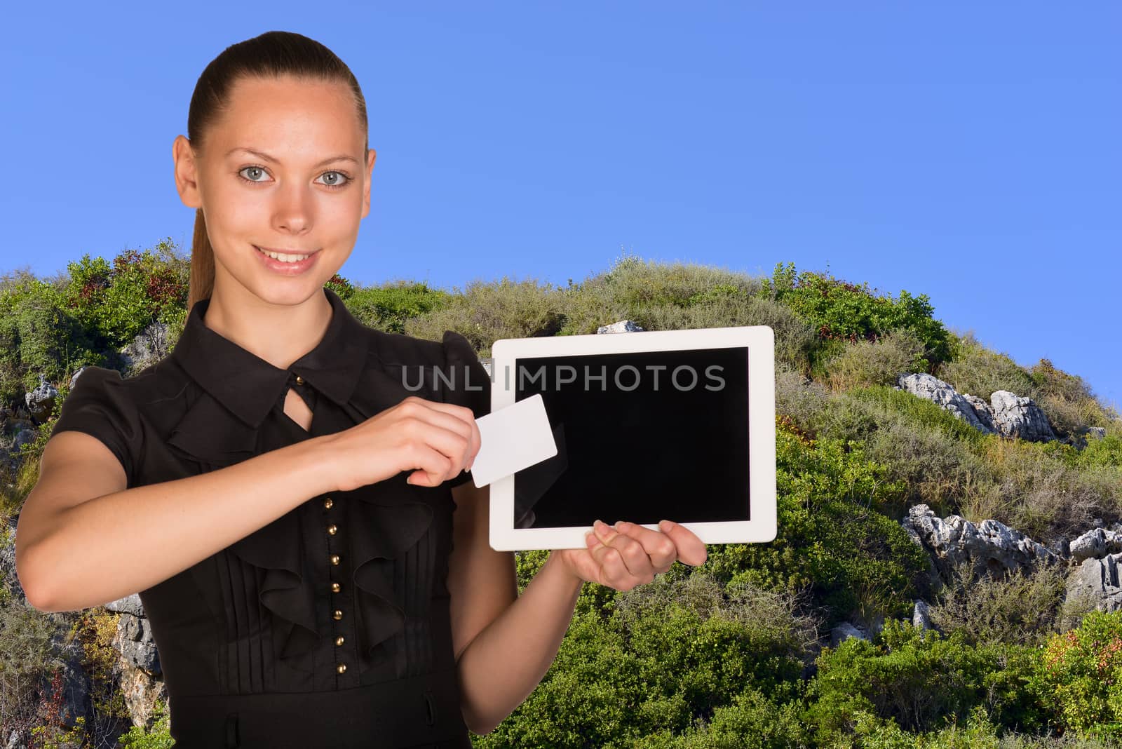 Beautiful businesswoman holding blank tablet PC and blank business card in front of PC screen. Blue sky and green hillside as backdrop