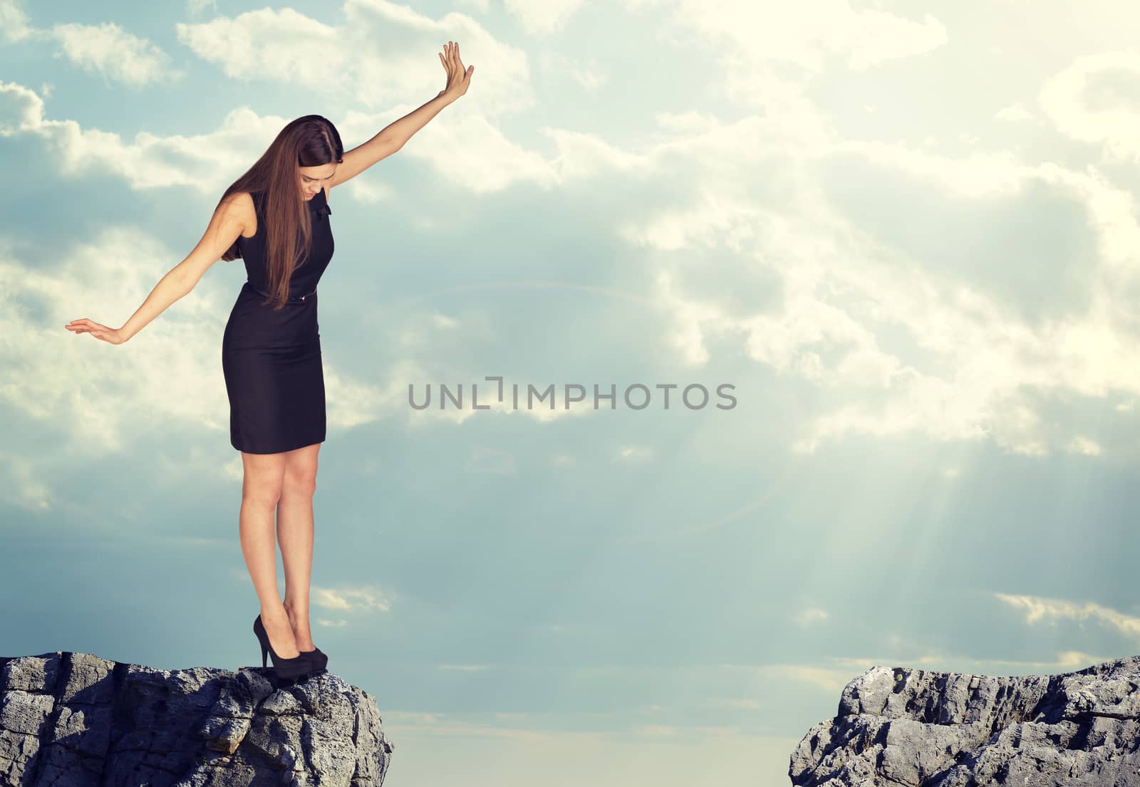 Businesswoman with her arms outstretched as for balance standing on the edge of rock gap and looking down into it. Sky and clouds as backdrop