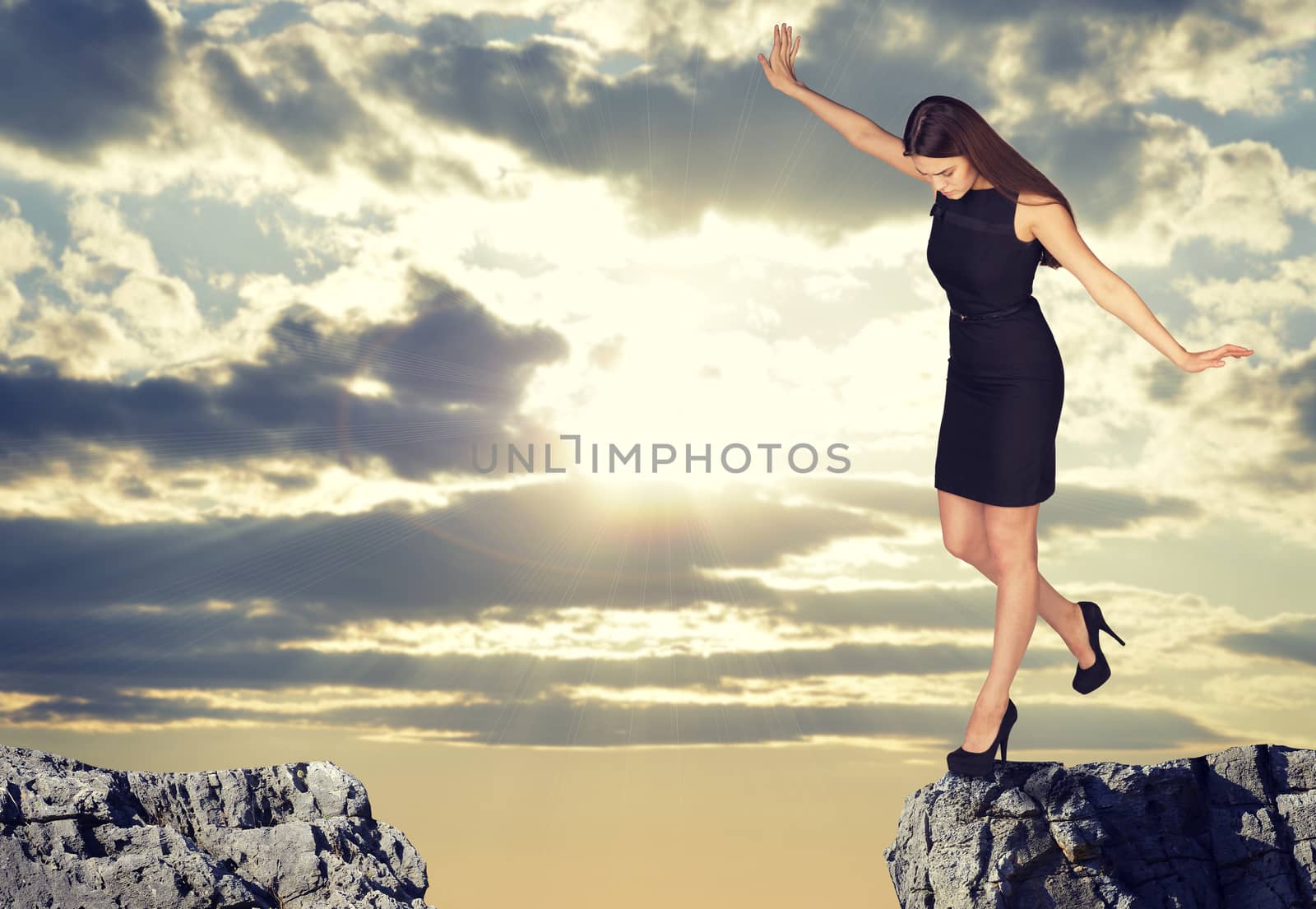 Businesswoman with her arms outstretched as for balance standing on the edge of rock gap and looking down into it. Sky with rays of sunshine through clouds as backdrop