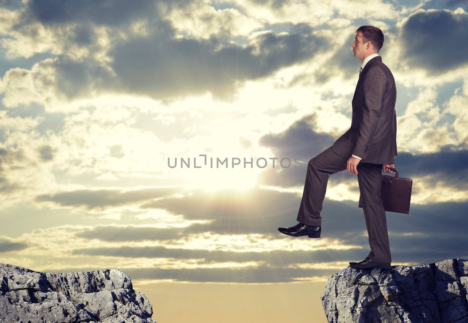 Businessman standing on the edge of rock gap, lifting his foot as if going to leap across it. Sky with sun shining through clouds as backdrop