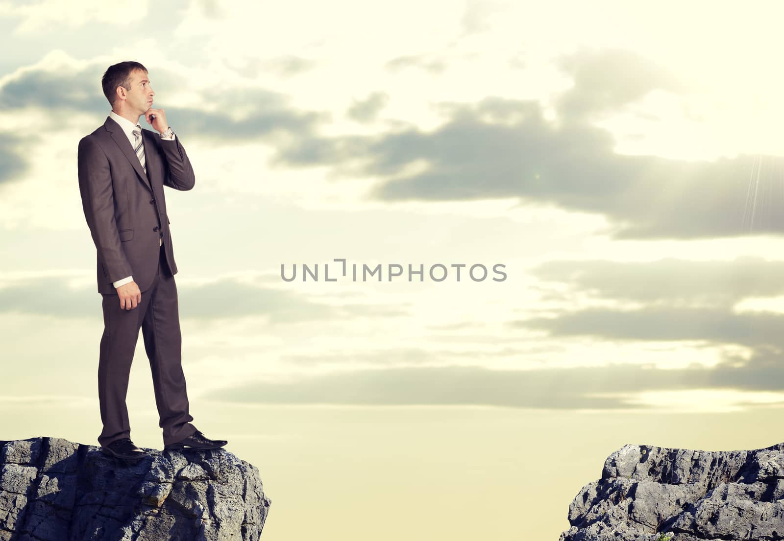 Businessman standing on the edge of rock gap in thoughtful pose. Sky and clouds as backdrop