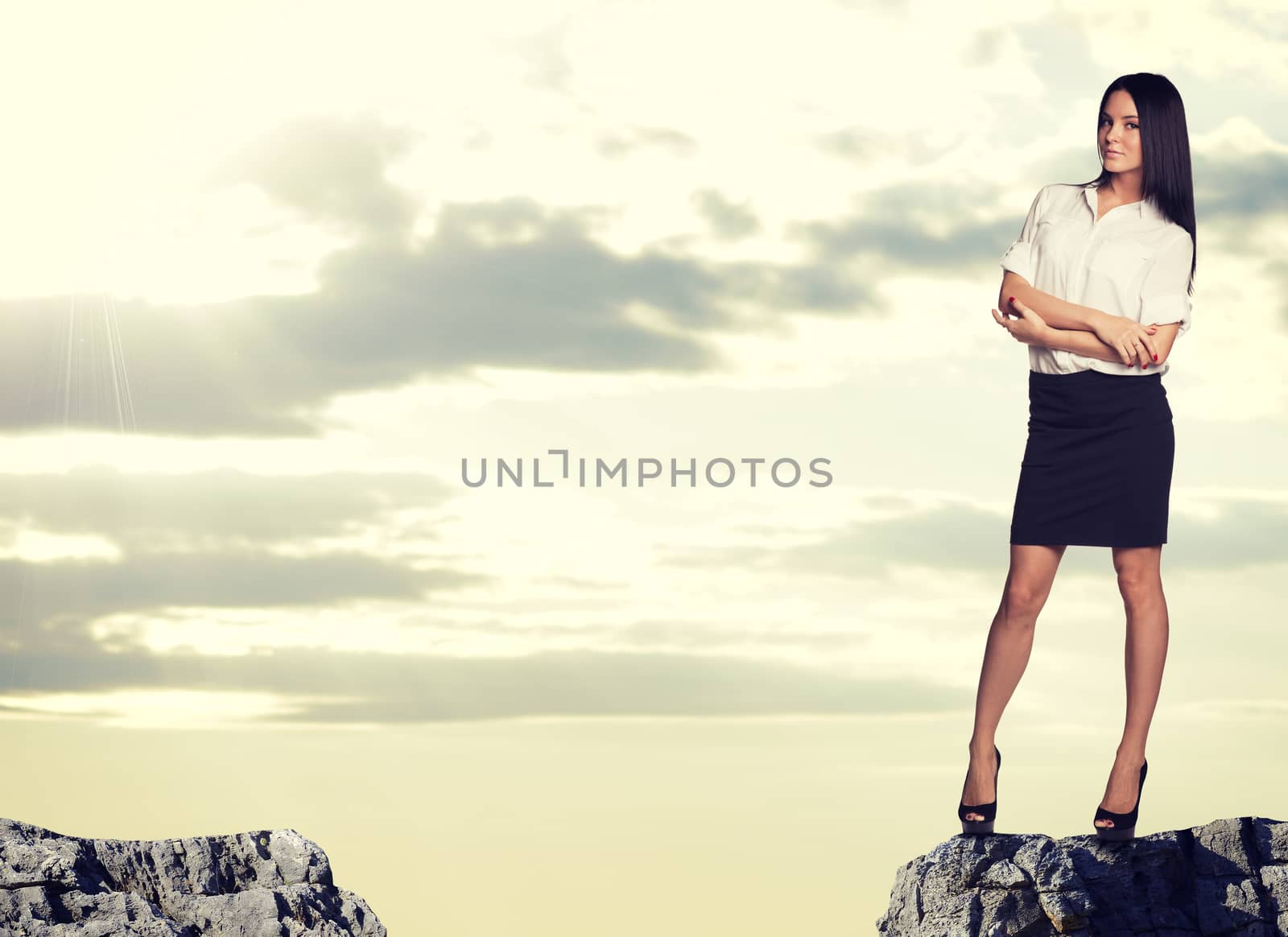 Businesswoman standing on the edge of rock gap. with her arms crossed on her breast, looking at camera, Sky and clouds as backdrop