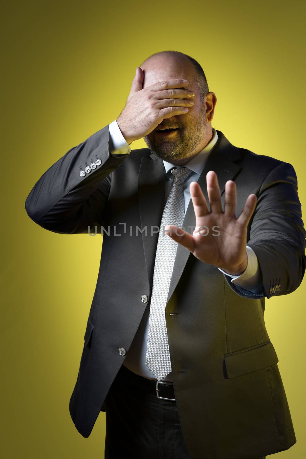 Stressed businessman in dark suit holding up hand in front of his eyes and parries with the other hand, against yellow background