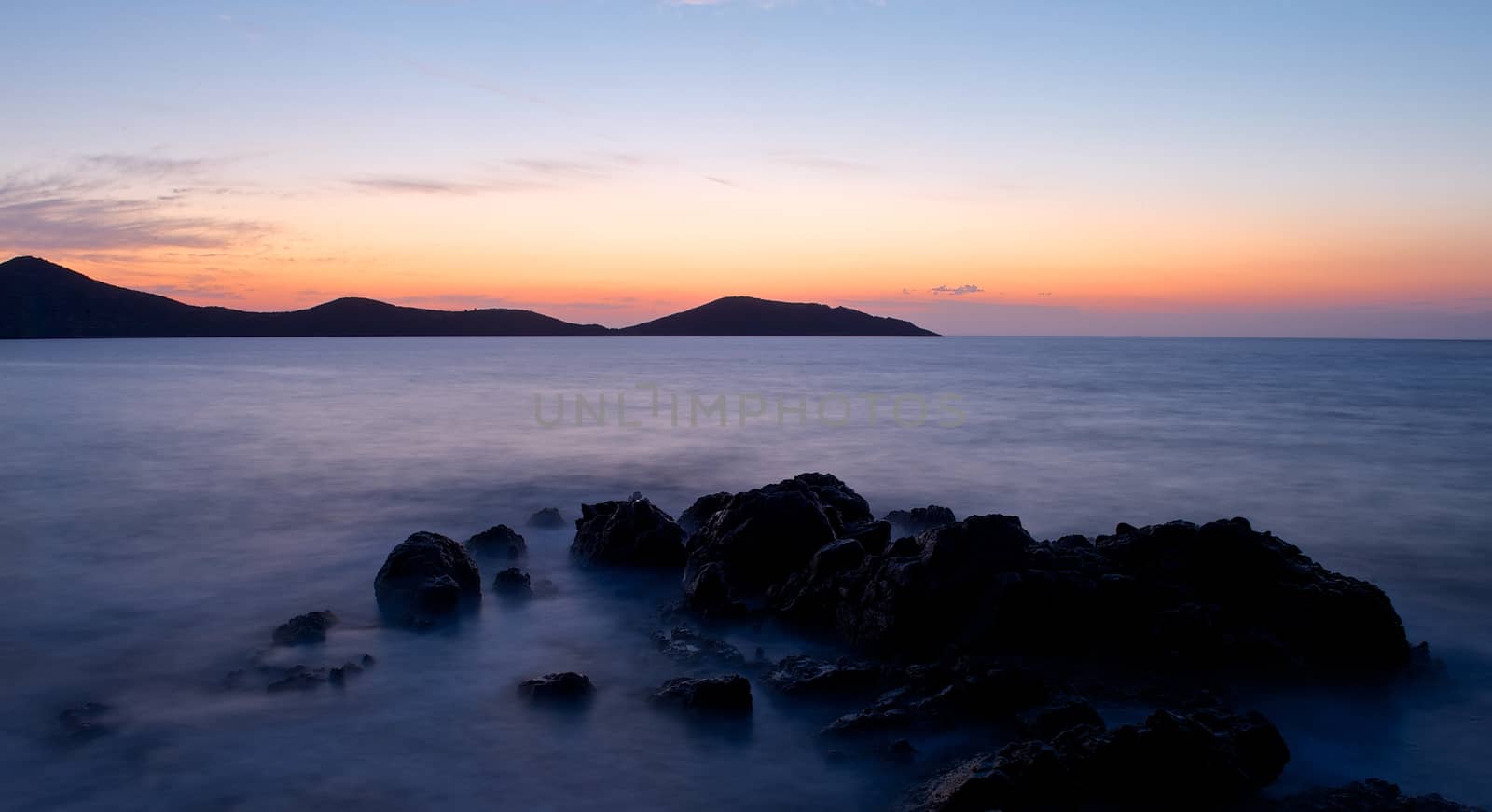 Horizontal sunset landscape of the island of Crete rocky fog covered seashore in the foreground and mountains in the back against golden hour sky.