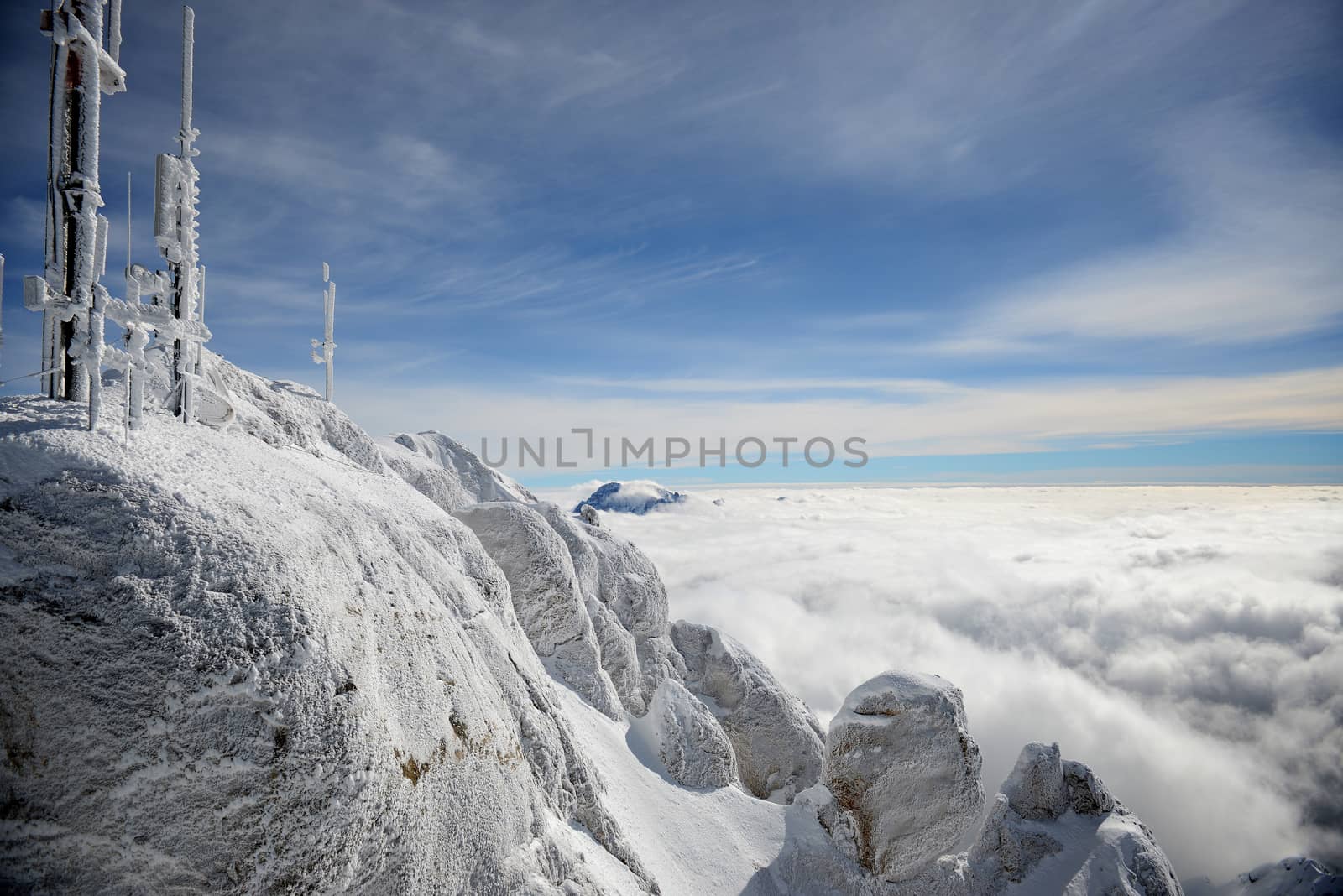 Horizontal landscape of a gray, colorless alpine mountaintop with frost covered radio antennae surrounded by a thick layer of clouds against vivid sky.