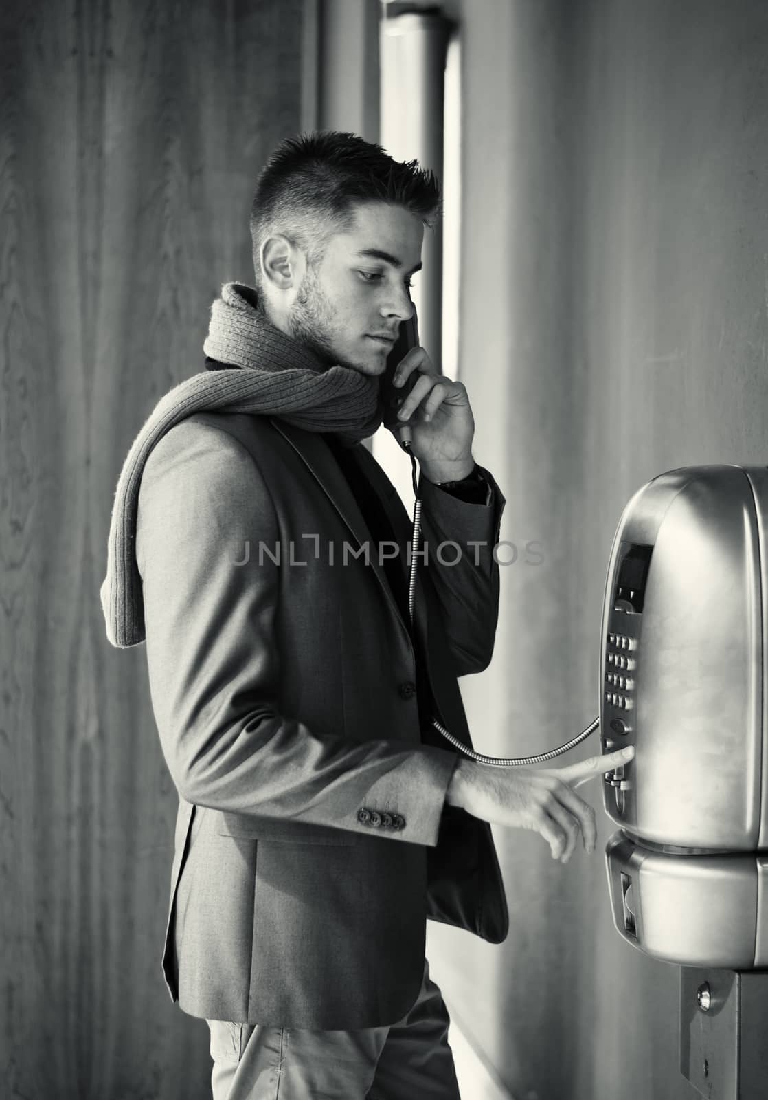 Handsome stylish young man in a tailored jacket and scarf standing using a pay phone waiting for a connection, profile view in greyscale