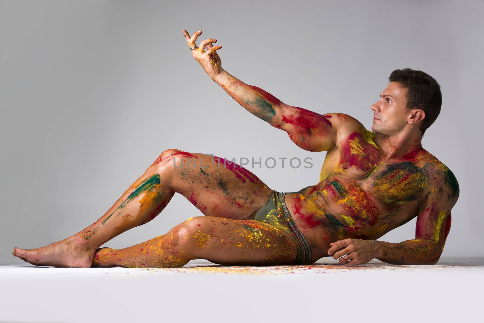 Muscular young man shirtless with skin painted with Holi colors, laying down on the floor striking a pose