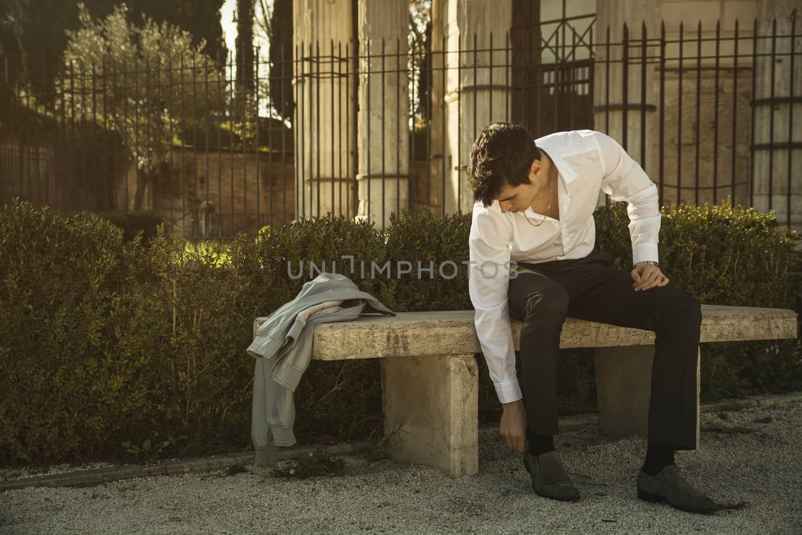 Handsome young man in European city, sitting on stone bench, adjusting a shoe