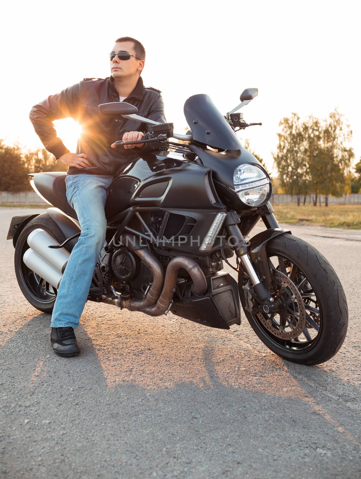 Biker man wearing a leather jacket and sunglasses sitting on his motorcycle outdoors