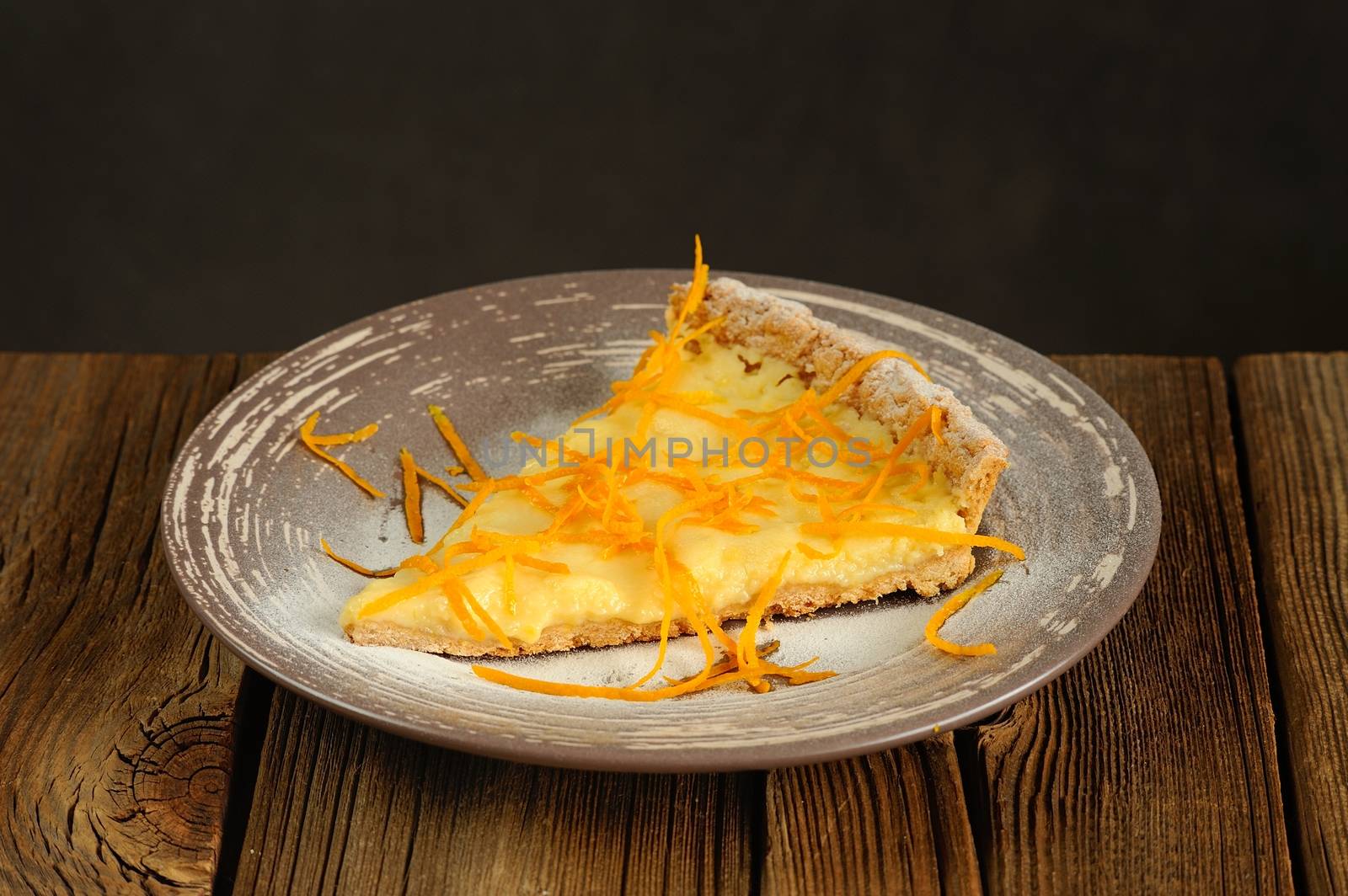 Piece on lemon tart with orange peel on wooden background