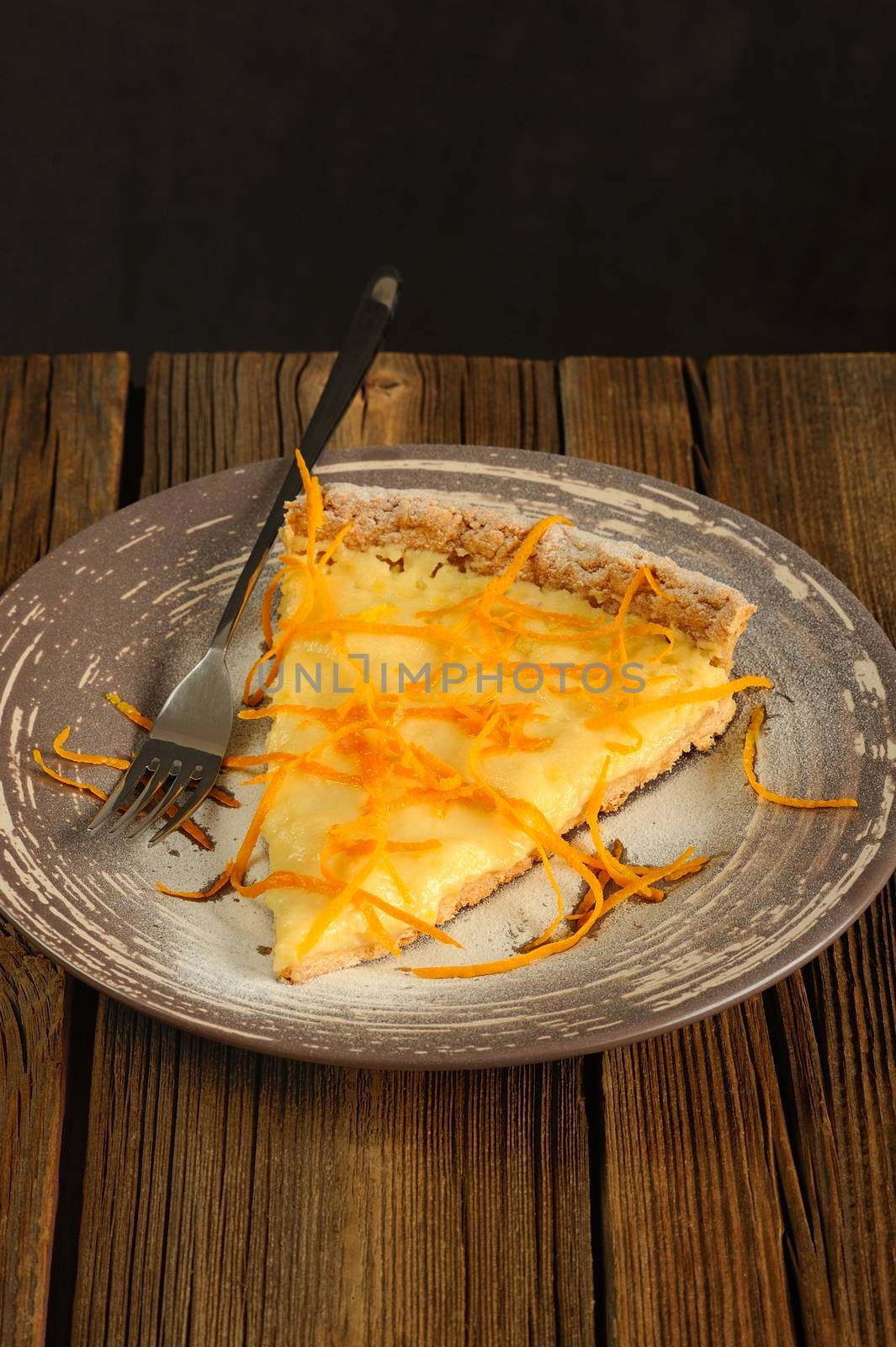 Piece on lemon tart with orange peel on wooden background