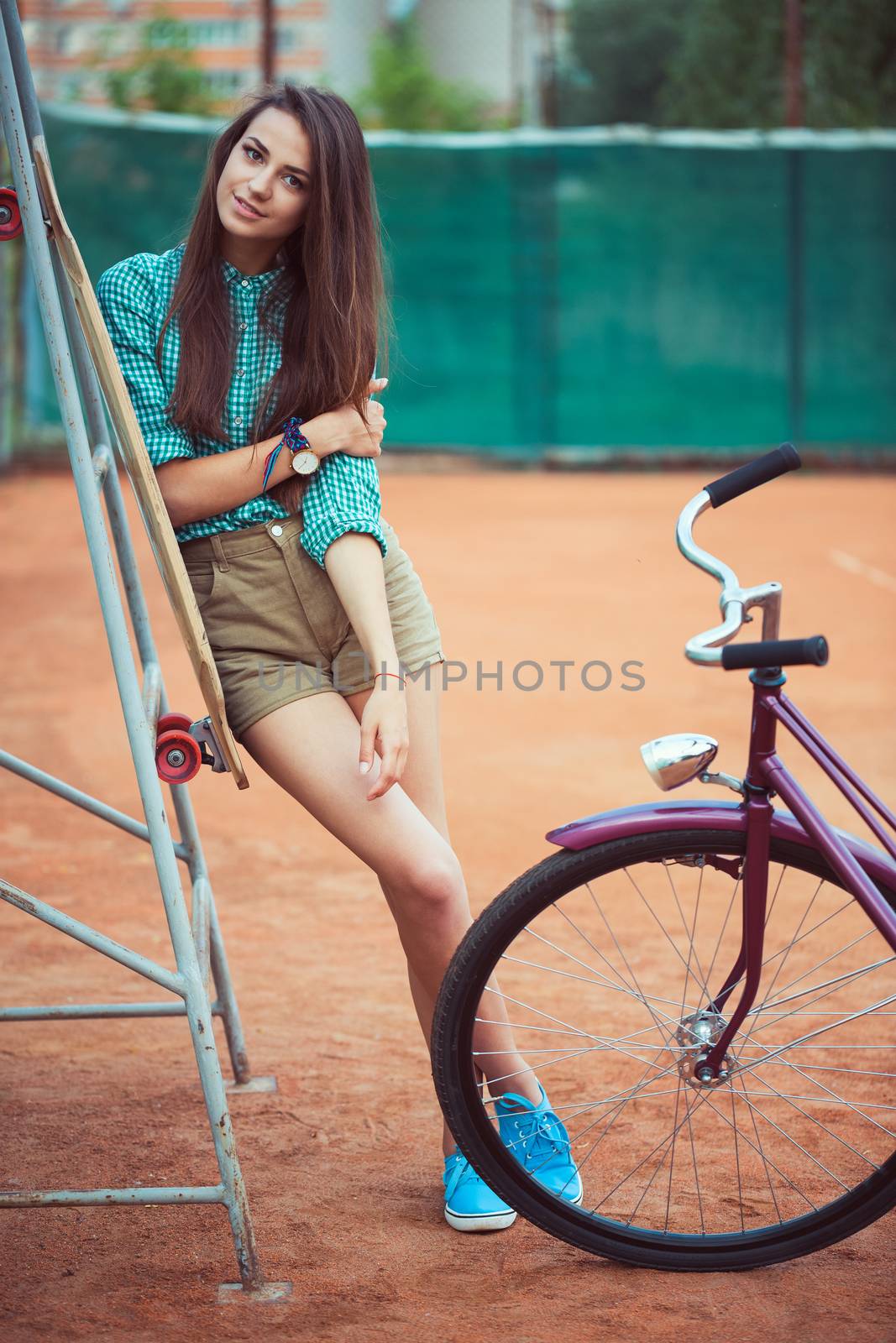 Beautiful young girl with longboard and bicycle standing on the tennis court, outdoor