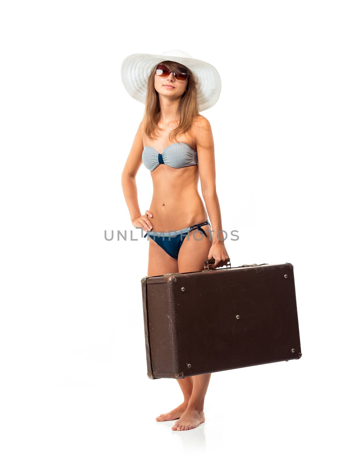 Full length portrait of a beautiful young woman posing in a bikini, hat and sunglasses with a suitcase in hand on white background