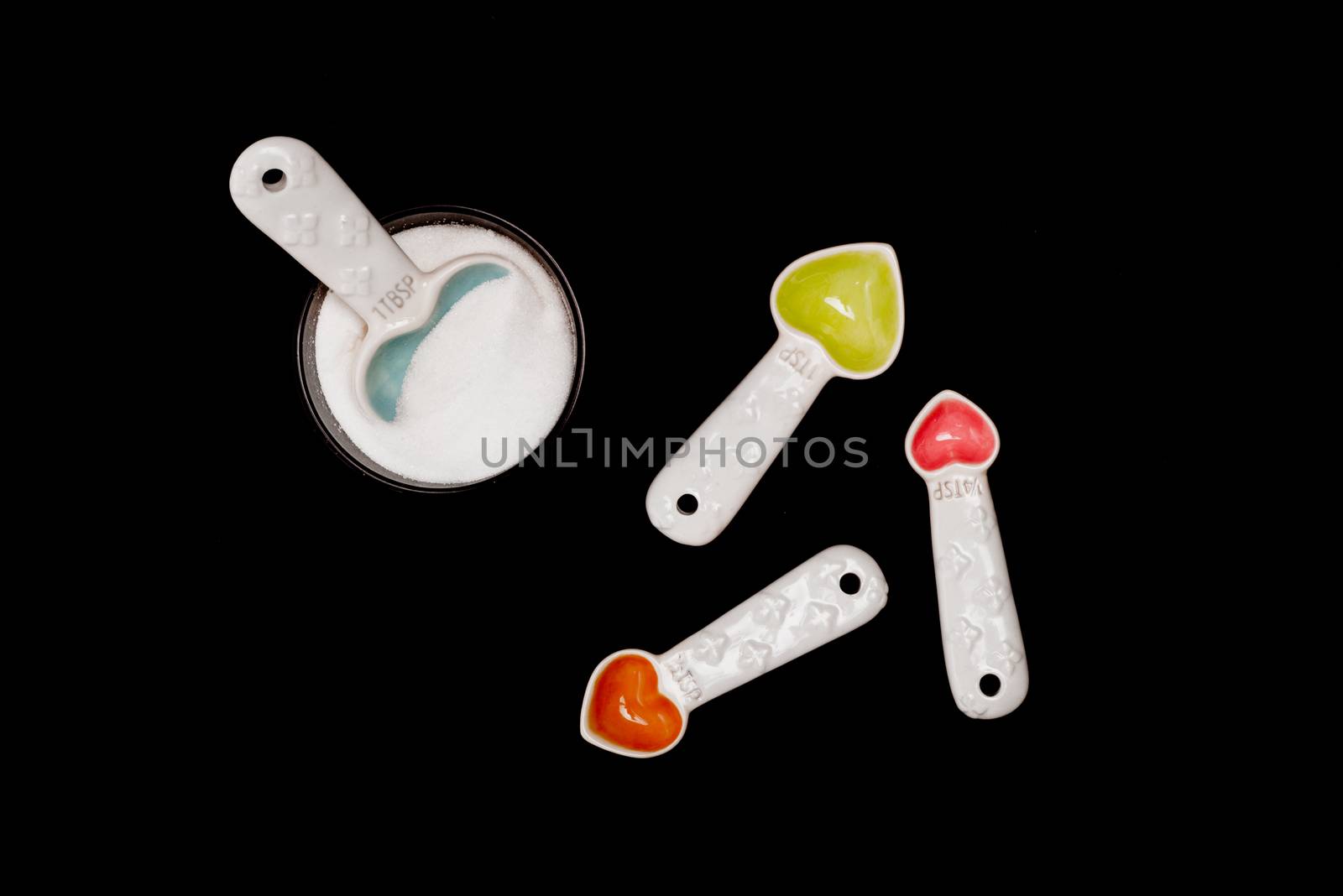 The largest of the heart shaped spoons is in the round bowl of sugar, while the others are facing in other directions on the isolated black backround.