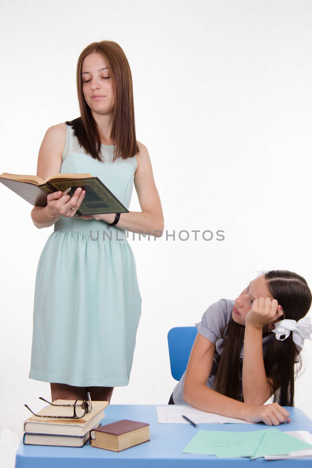 Teacher standing at desk behind which sits a tired student