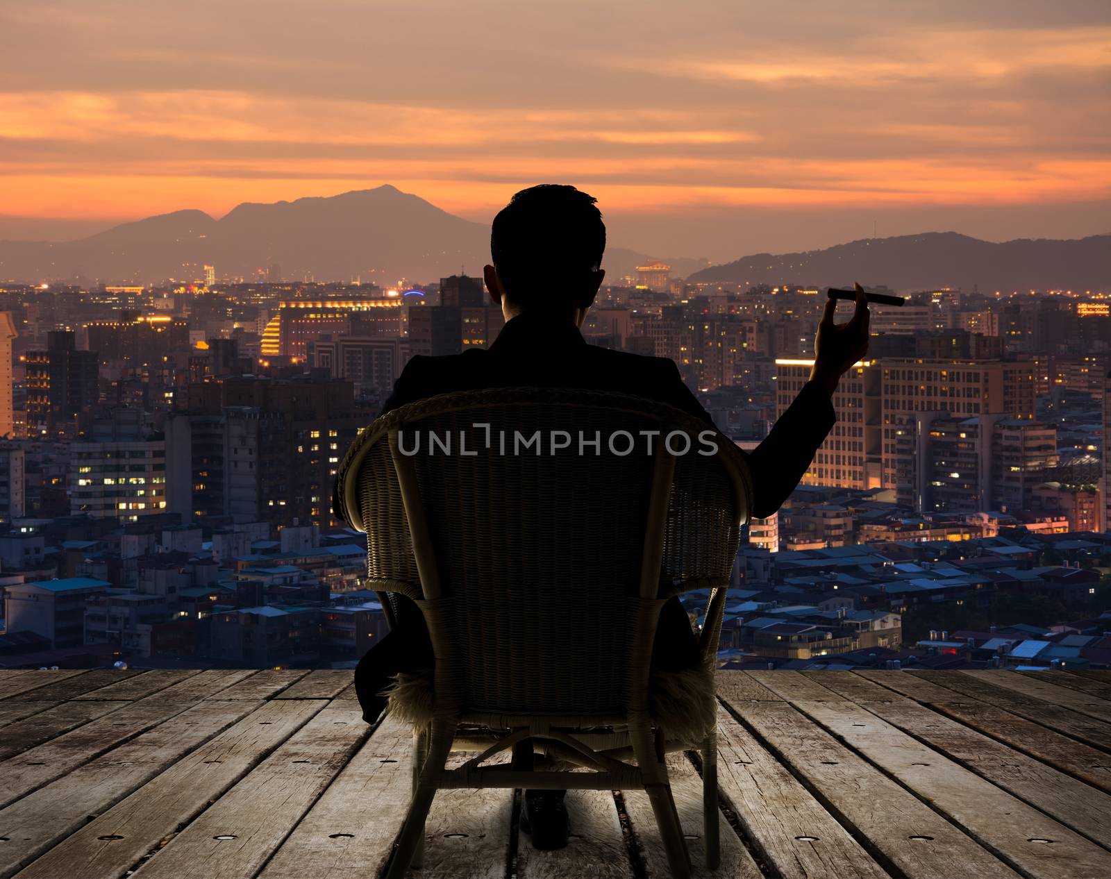 Silhouette of businessman sit on chair and hold a cigar and looking at the city in night.