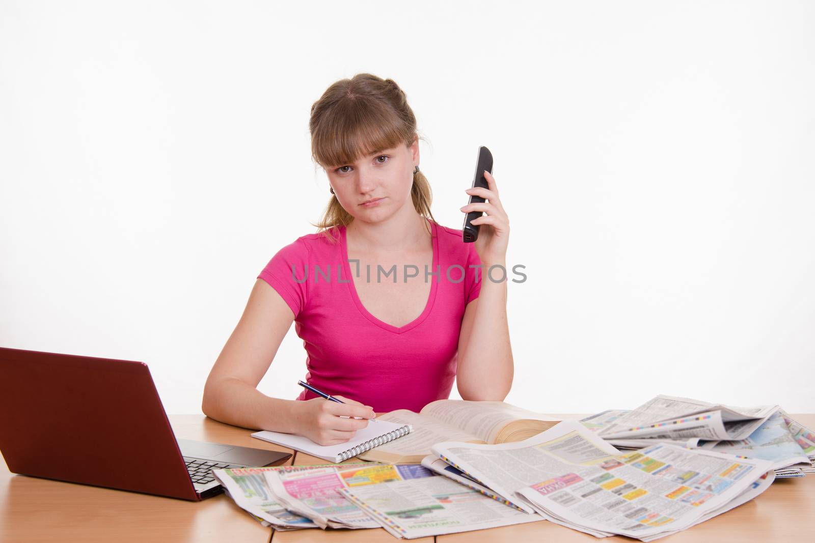 A young girl sits at a table with a pile of newspapers, encyclopedias and a laptop