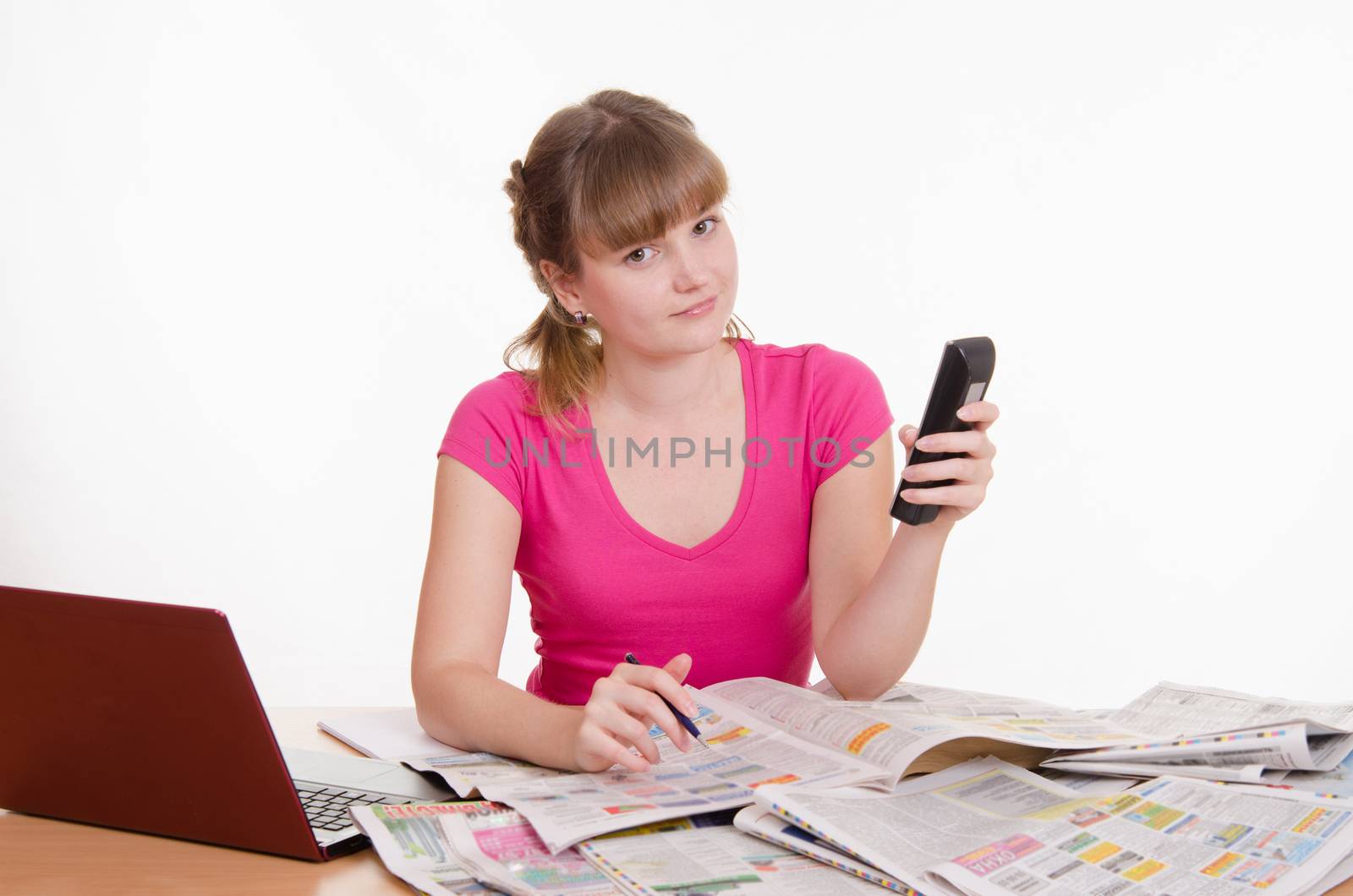 A young girl sits at a table with a pile of newspapers, encyclopedias and a laptop
