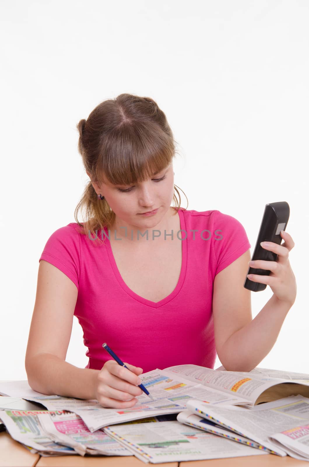 A young girl sits at a table with a pile of newspapers, encyclopedias and a laptop