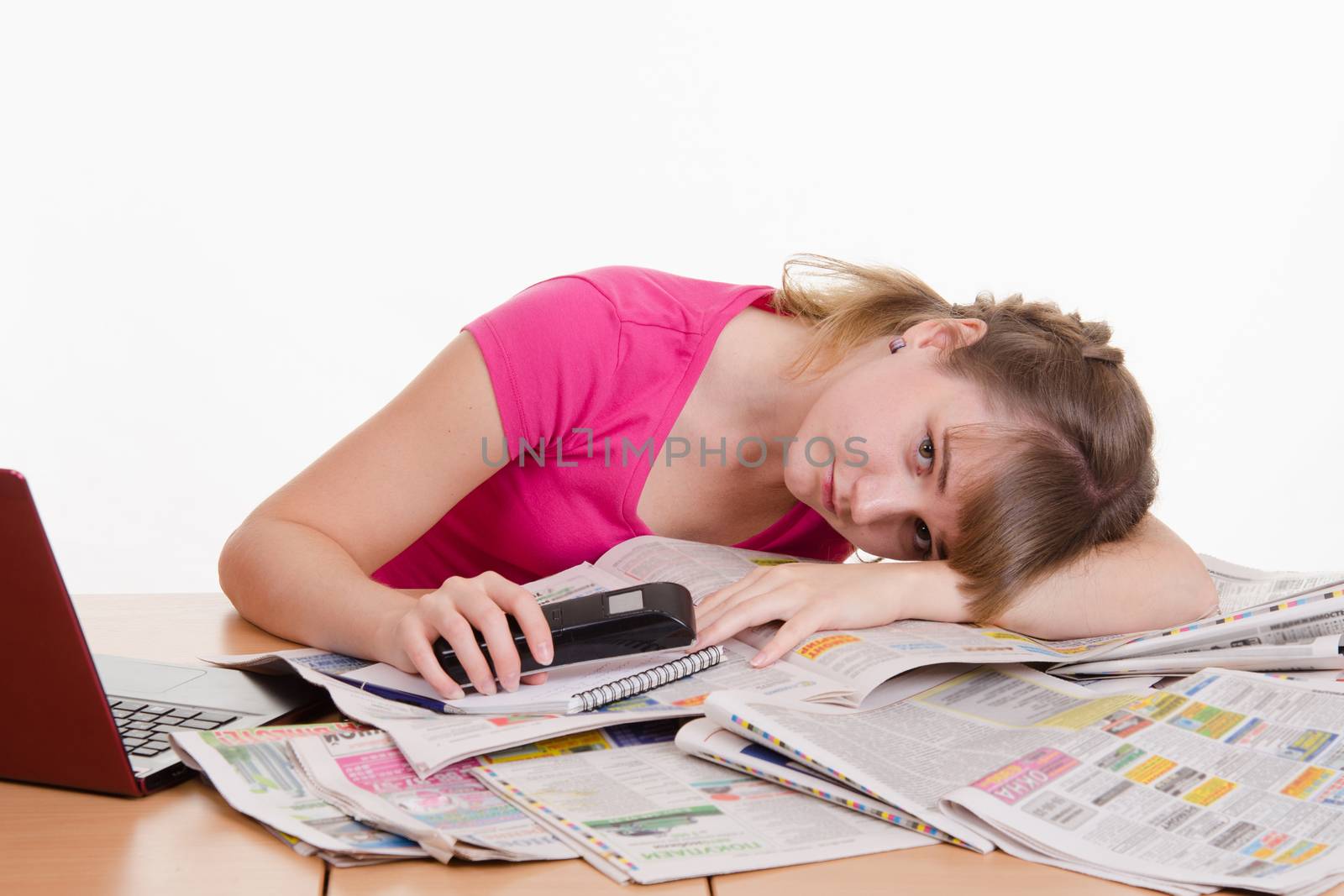 A young girl sits at a table with a pile of newspapers, encyclopedias and a laptop
