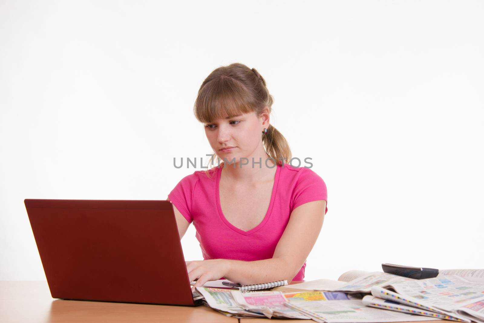A young girl sits at a table with a pile of newspapers, encyclopedias and a laptop