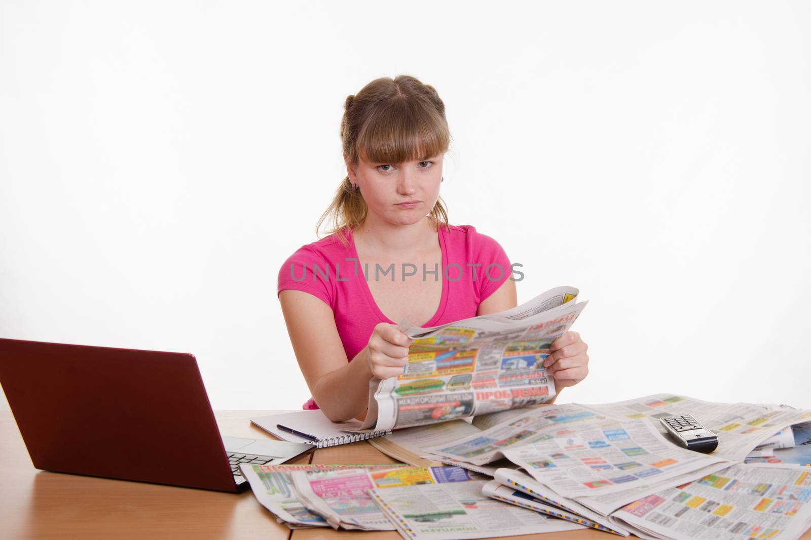 A young girl sits at a table with a pile of newspapers, encyclopedias and a laptop