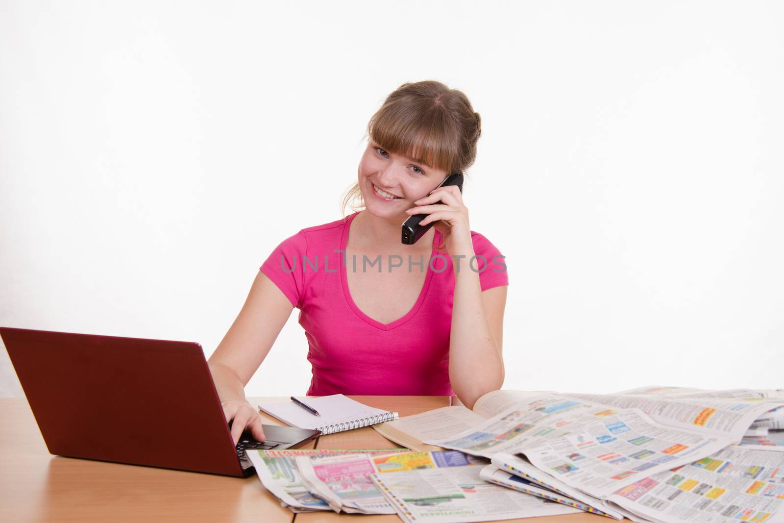 A young girl sits at a table with a pile of newspapers, encyclopedias and a laptop