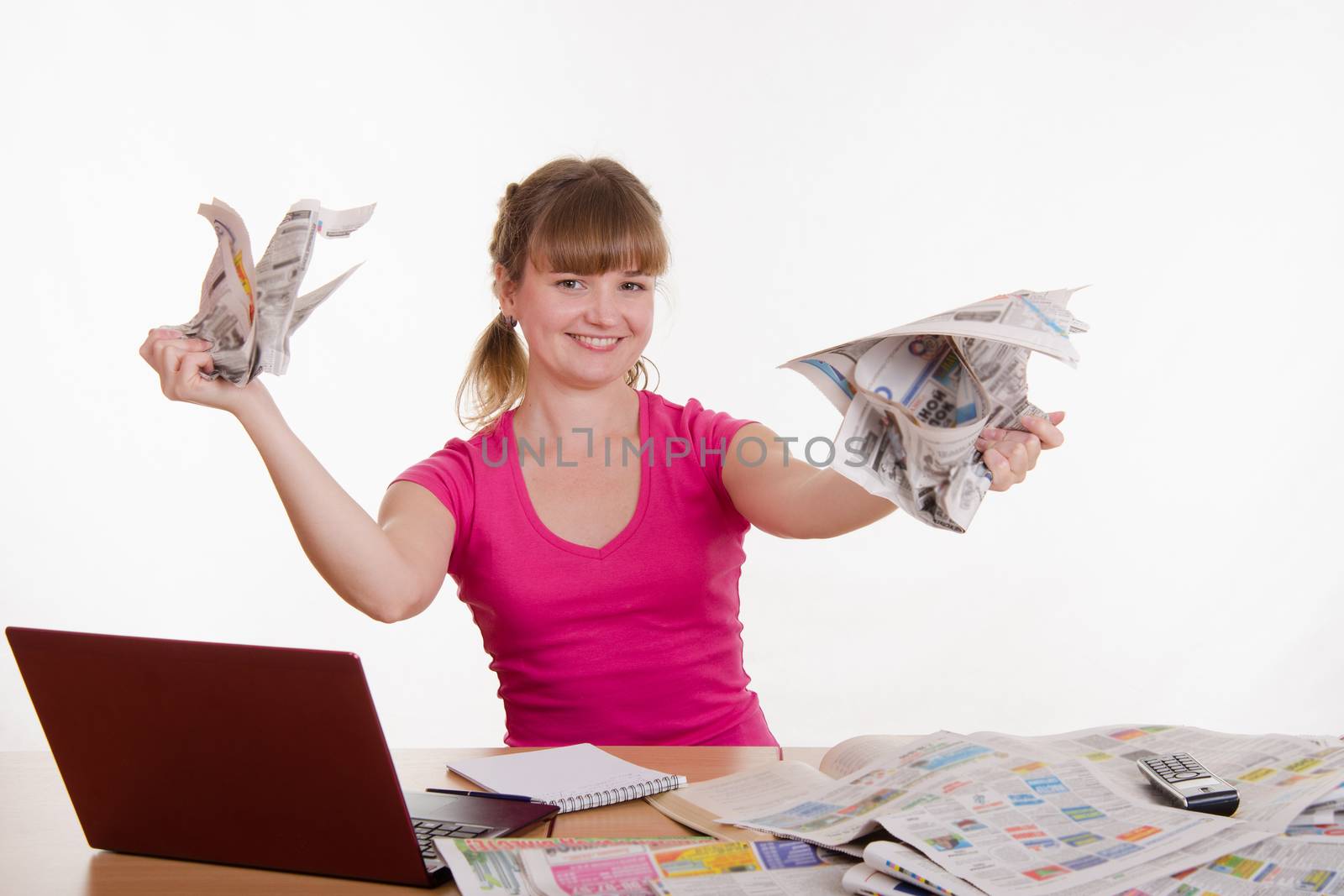 A young girl sits at a table with a pile of newspapers, encyclopedias and a laptop