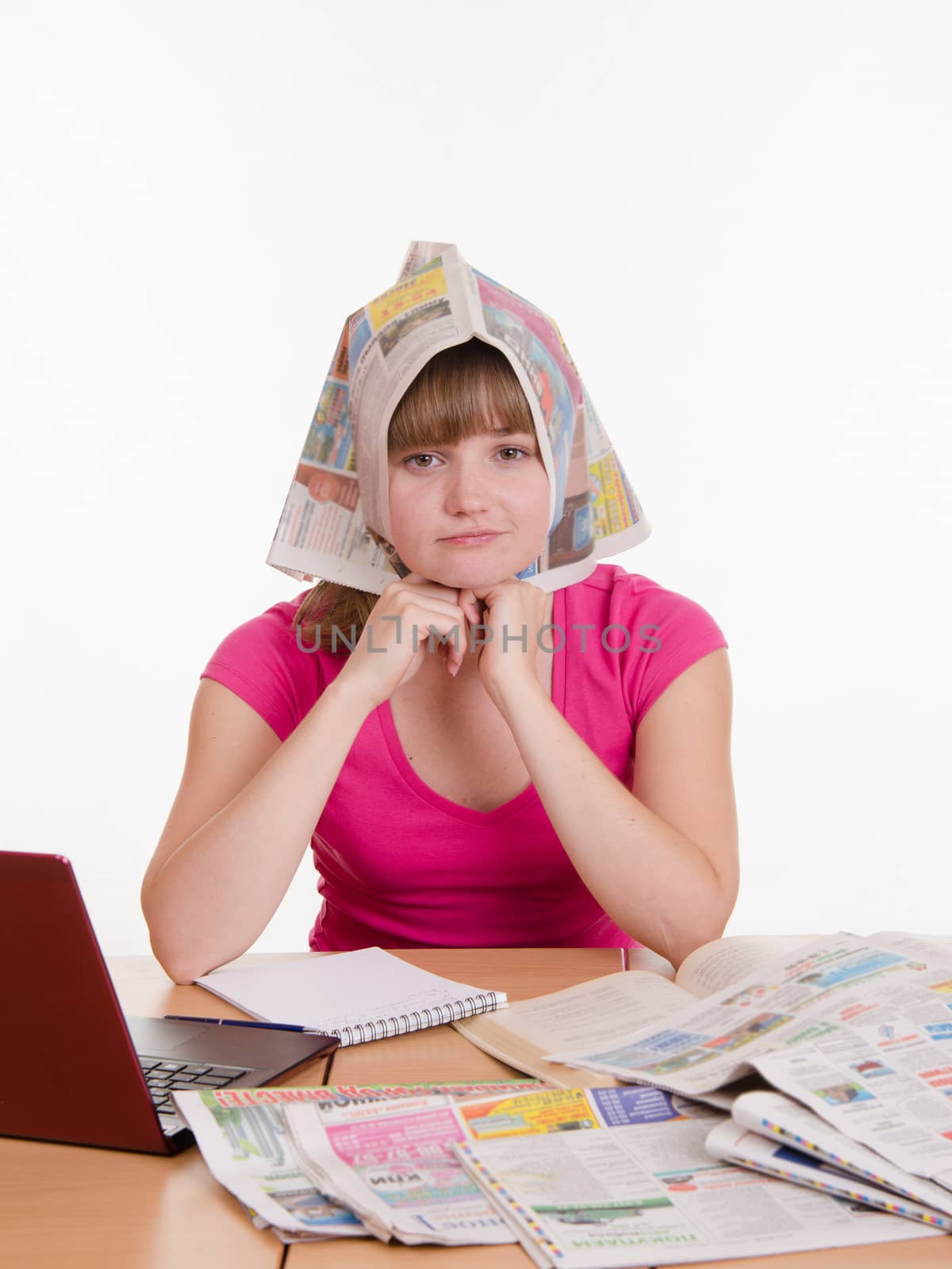 A young girl sits at a table with a pile of newspapers, encyclopedias and a laptop