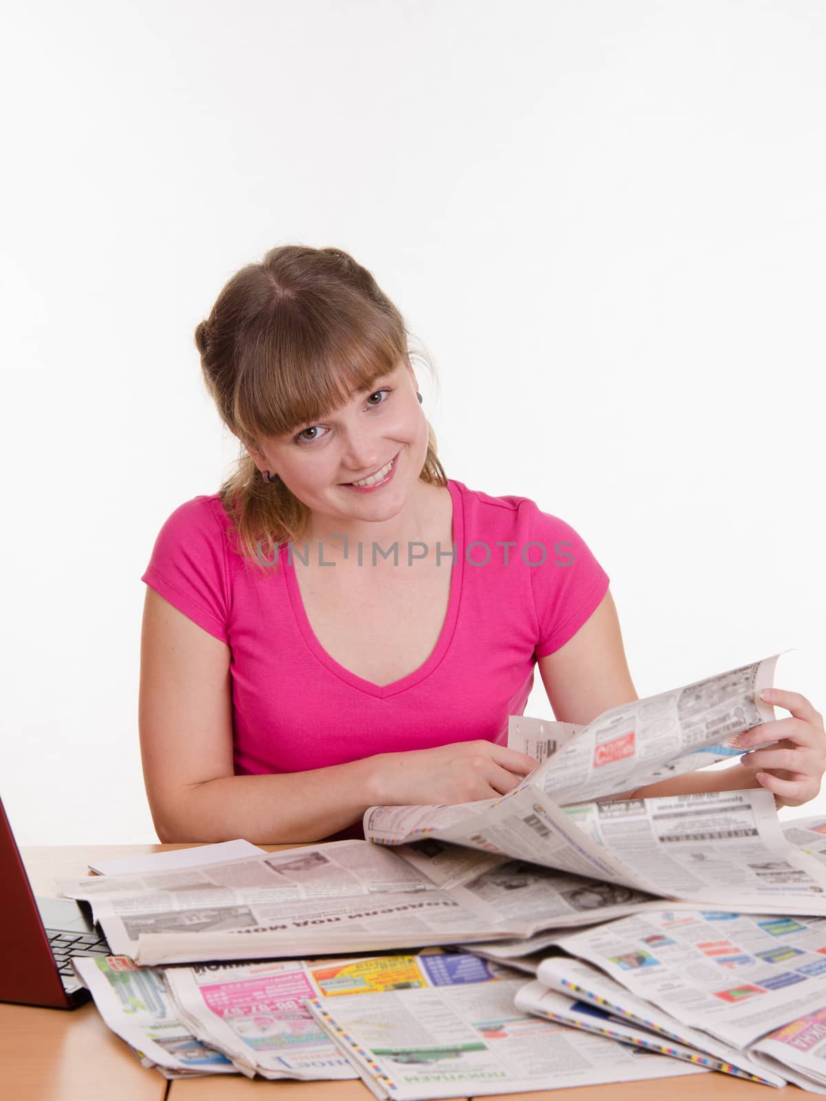 A young girl sits at a table with a pile of newspapers, encyclopedias and a laptop