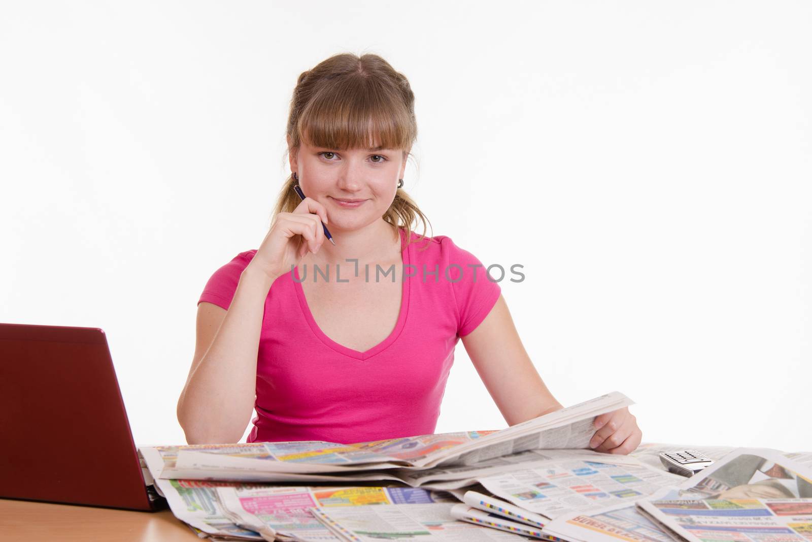 A young girl sits at a table with a pile of newspapers, encyclopedias and a laptop