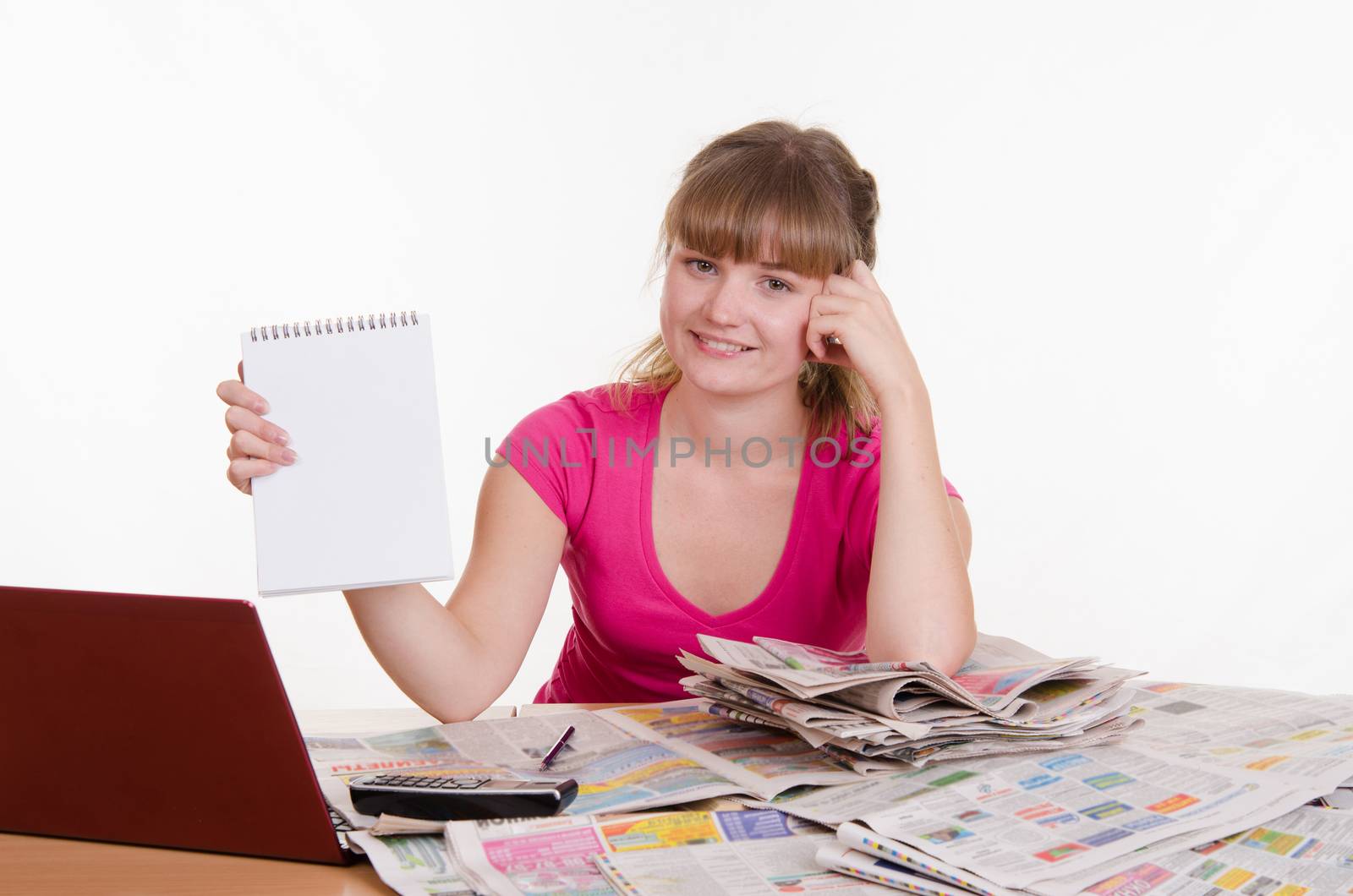 A young girl sits at a table with a pile of newspapers, encyclopedias and a laptop