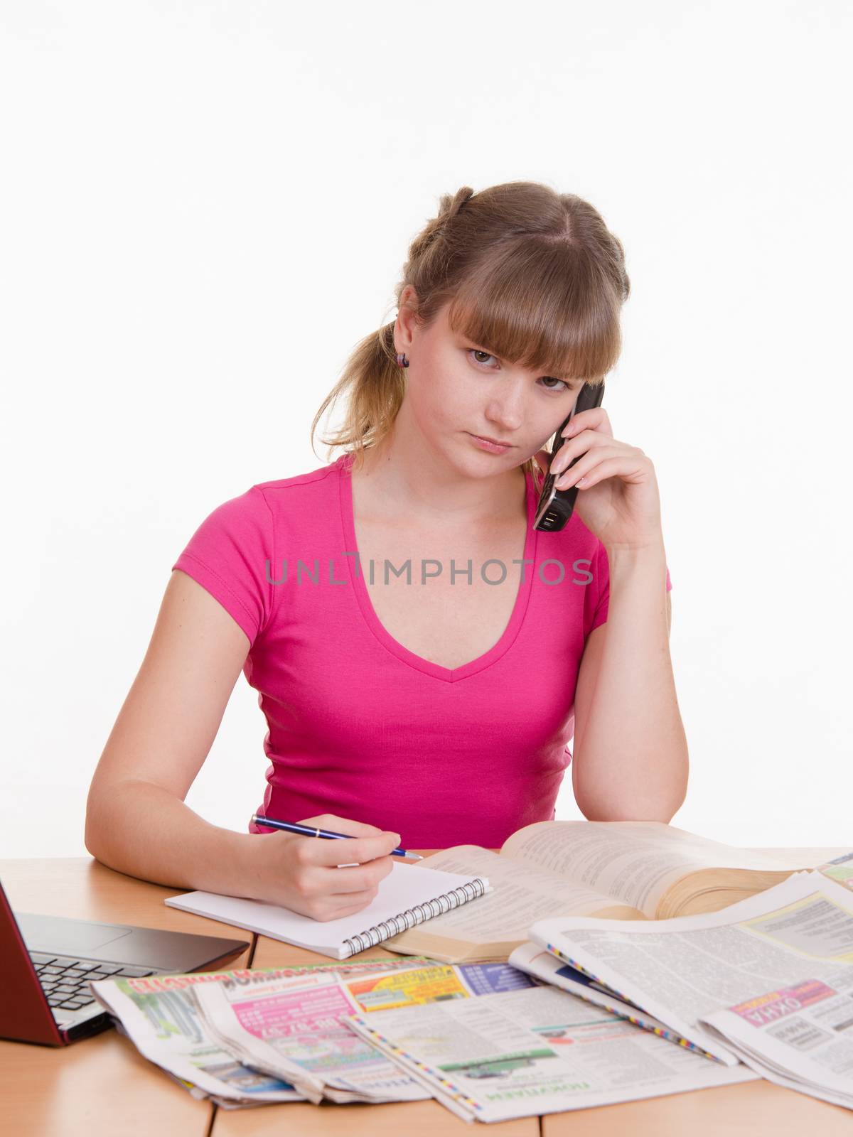 A young girl sits at a table with a pile of newspapers, encyclopedias and a laptop
