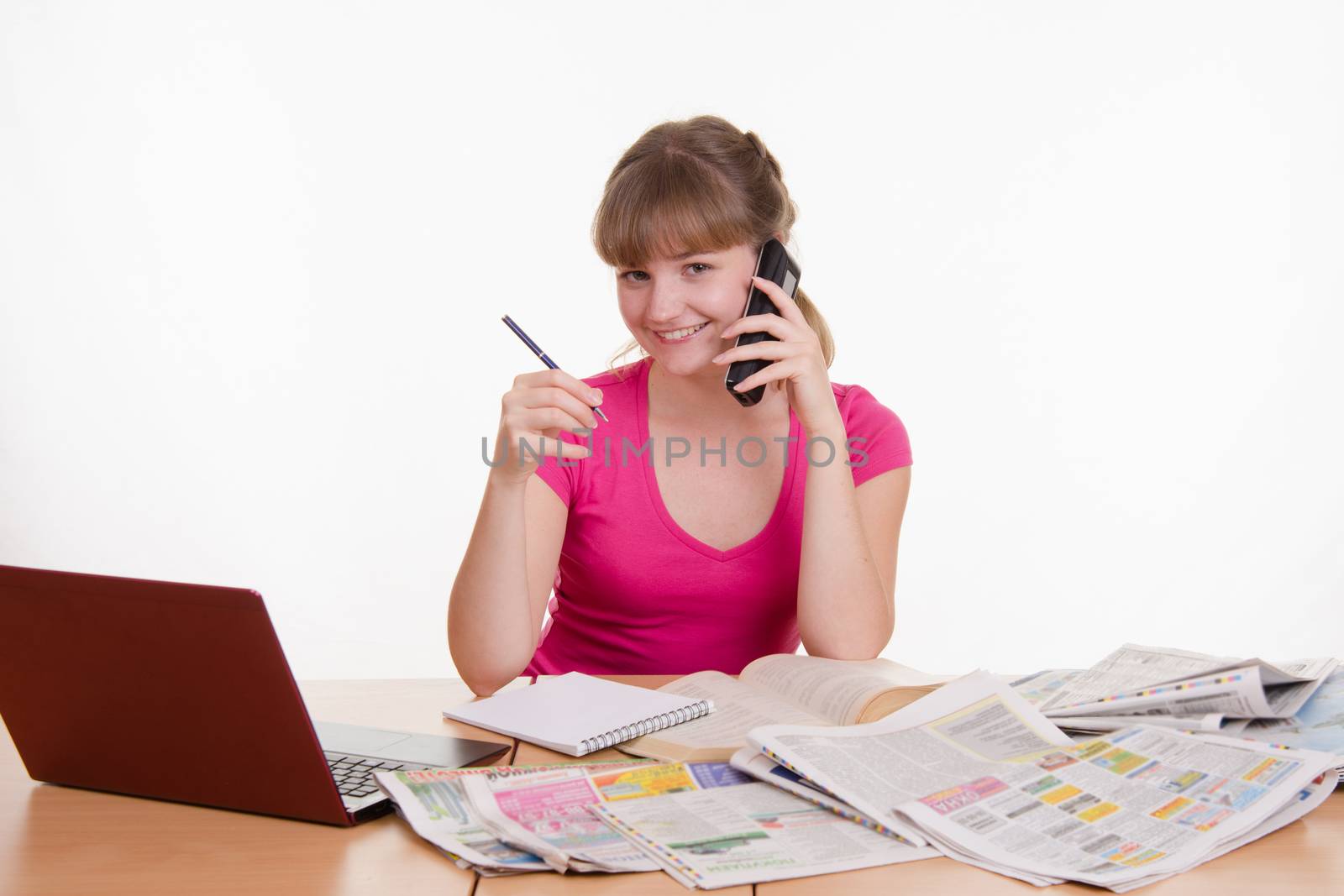 A young girl sits at a table with a pile of newspapers, encyclopedias and a laptop