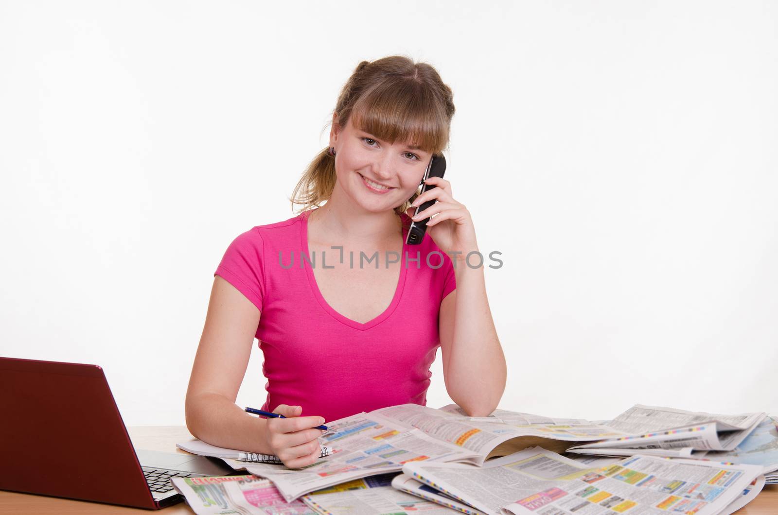 A young girl sits at a table with a pile of newspapers, encyclopedias and a laptop