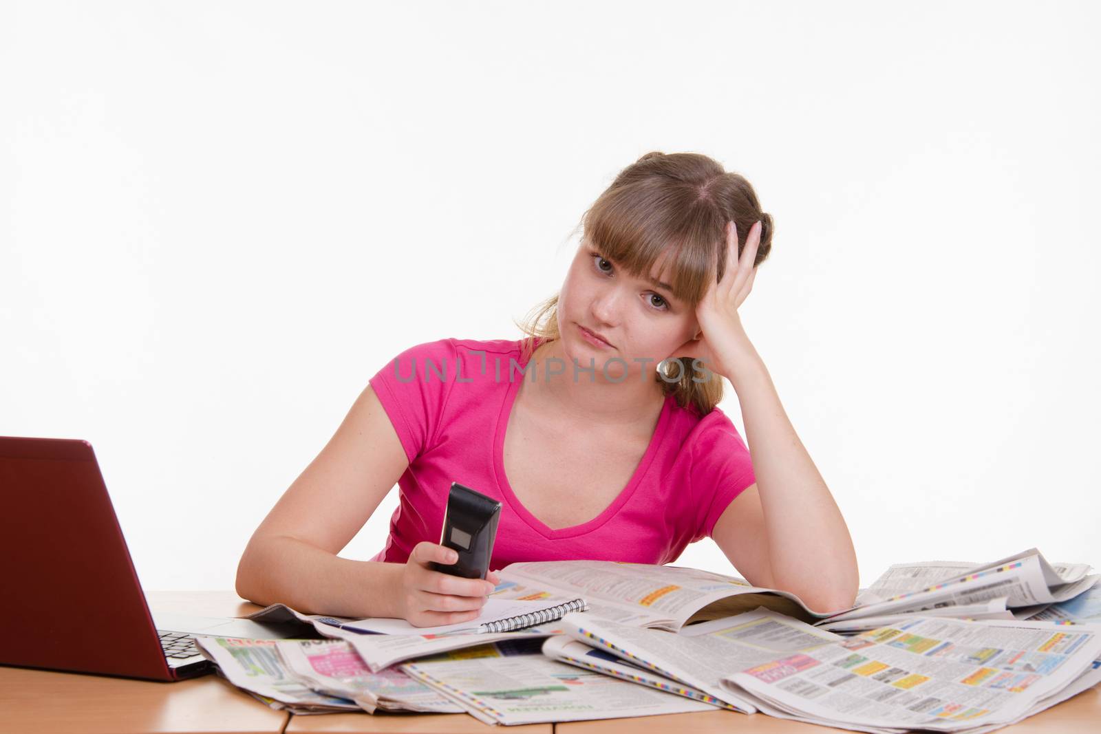 A young girl sits at a table with a pile of newspapers, encyclopedias and a laptop