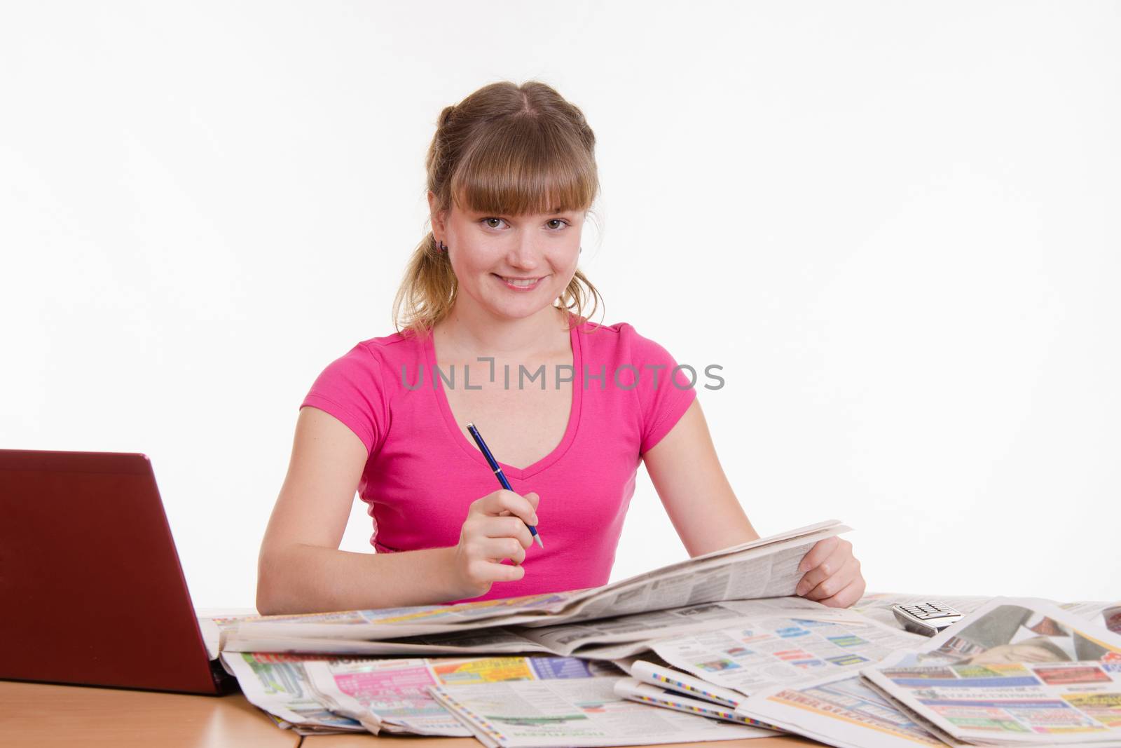 A young girl sits at a table with a pile of newspapers, encyclopedias and a laptop