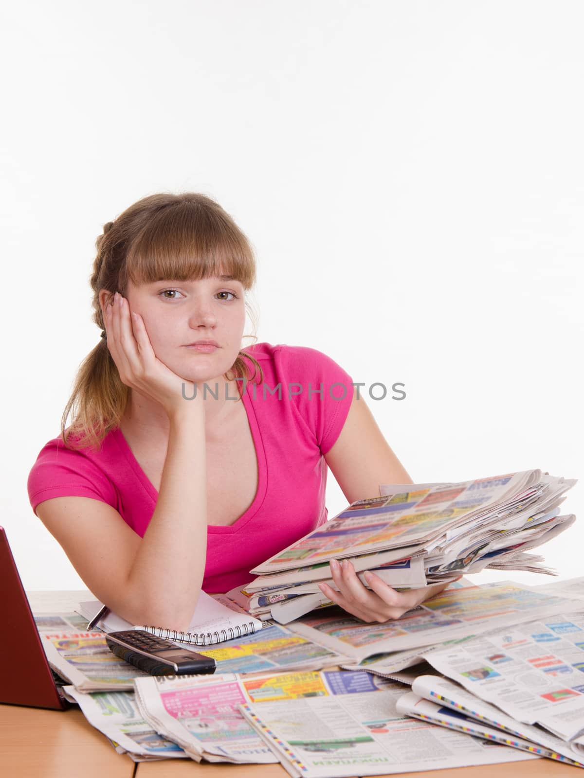 A young girl sits at a table with a pile of newspapers, encyclopedias and a laptop