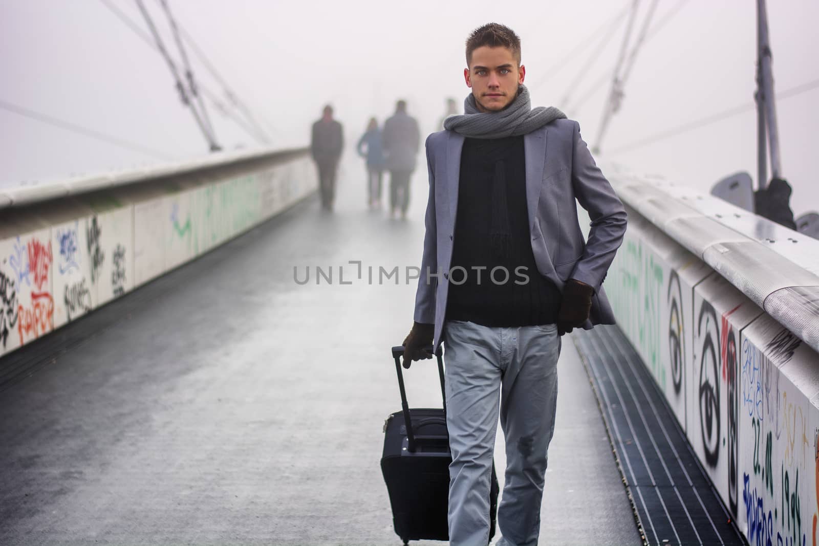 Trendy handsome man walking across a bridge on a misty cold day pulling a suitcase behind him as he departs on a trip