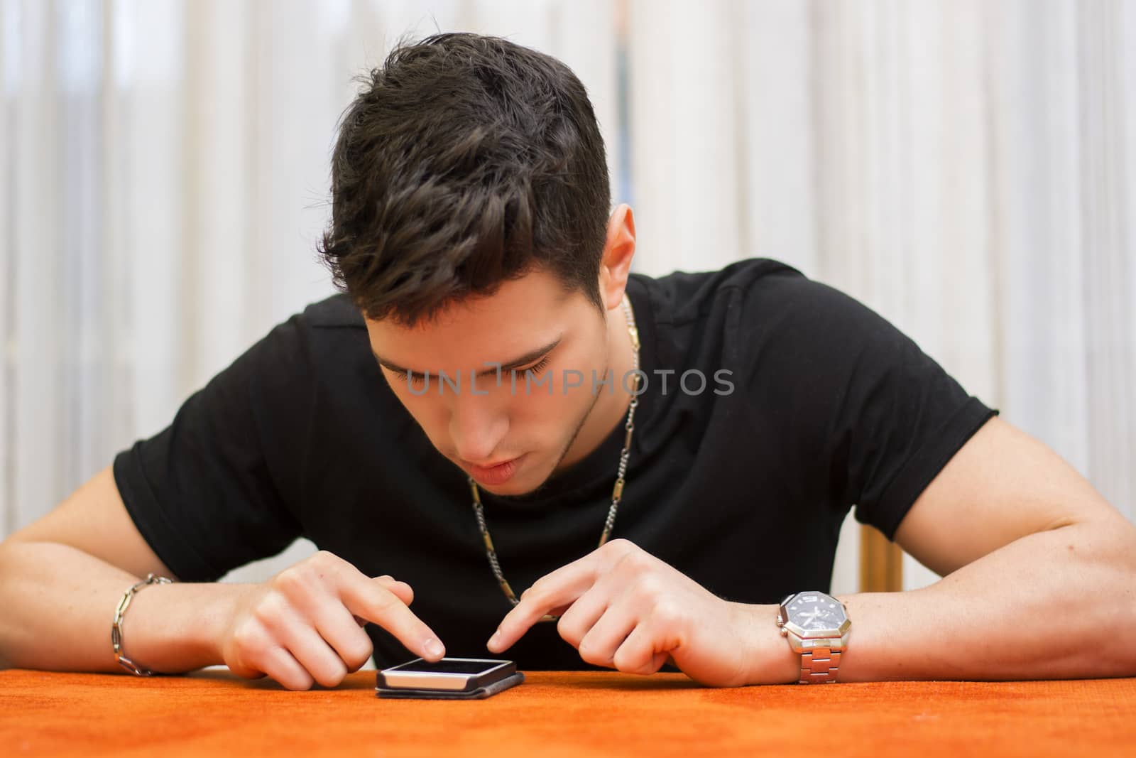 Young man sitting at a table in his home typing a text message on his phone with his fingers as he communicates with friends