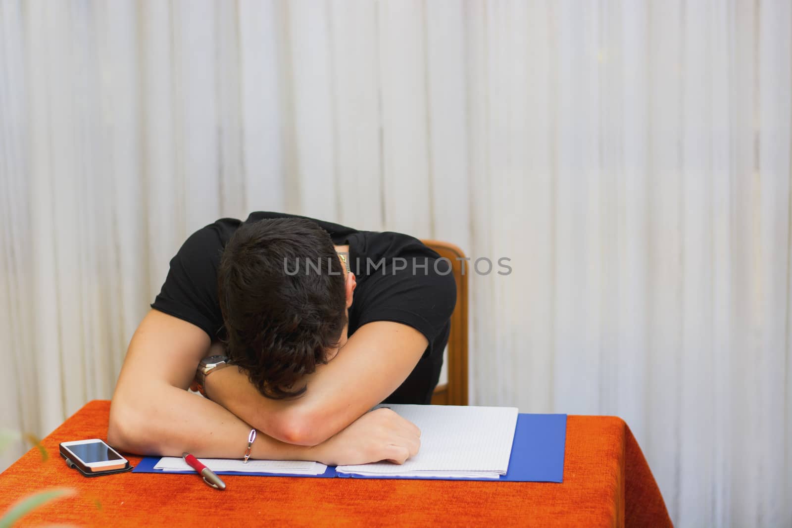 Tired, exhausted or despondent young man sitting at a table with a folder of paperwork leaning forwards with his head on his arms