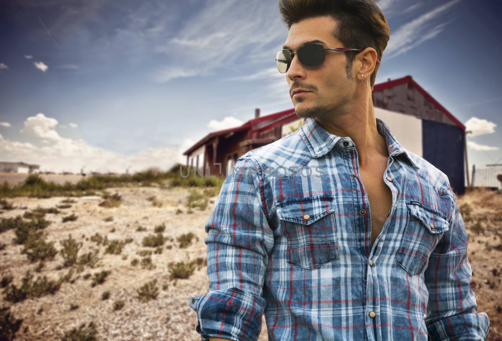 Handsome fashionable man in sunglasses and a blue checked shirt posing outdoors with a wooden cabin behind and copyspace looking into the centre of the frame