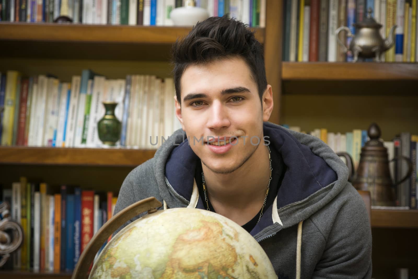 Handsome young man holding a globe indoors at home standing smiling at the camera in front of a bookcase filled with books in a conceptual image