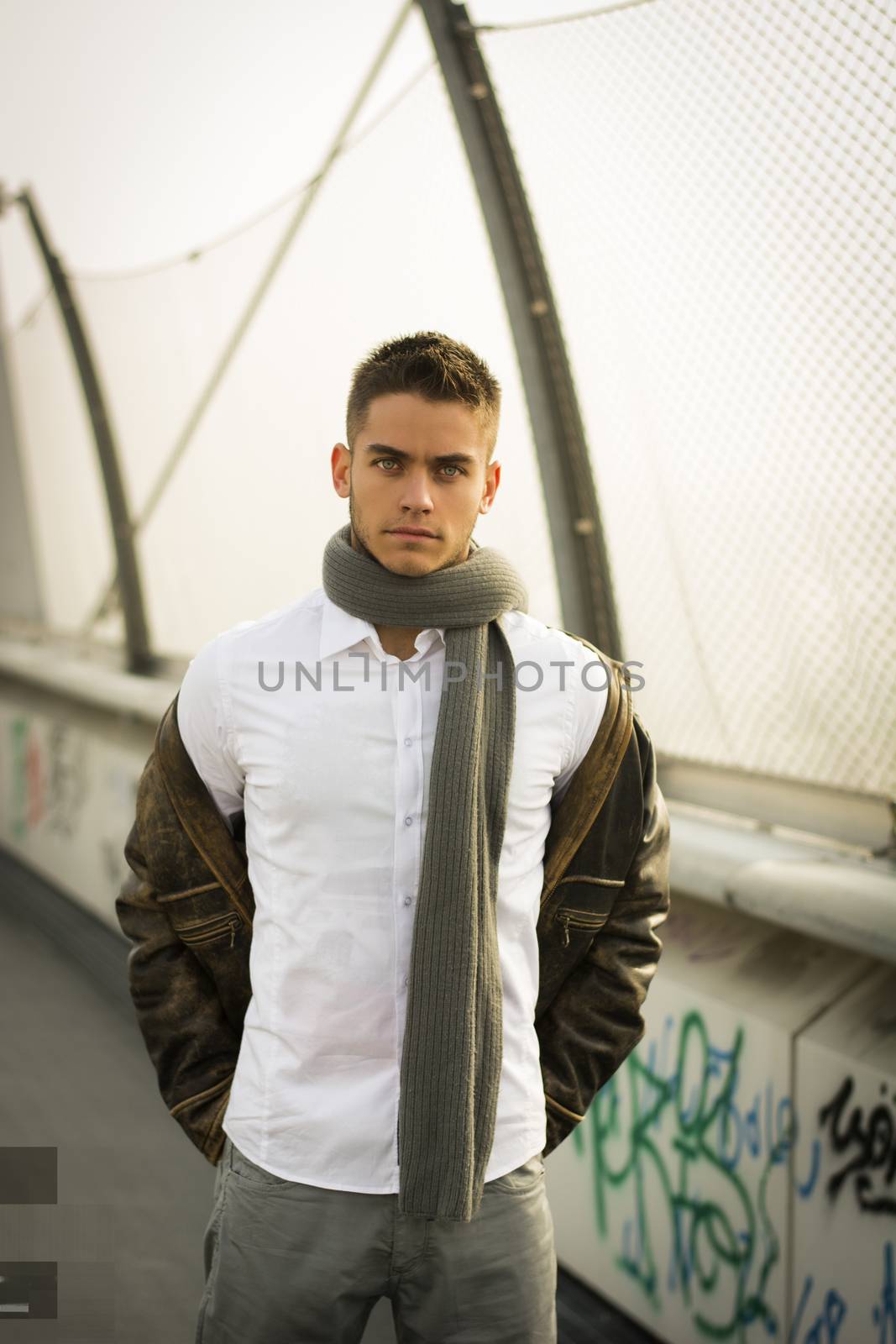 Handsome trendy young man standing on a sidewalk wearing a fashionable jacket and scarf in a relaxed confident pose looking at the camera